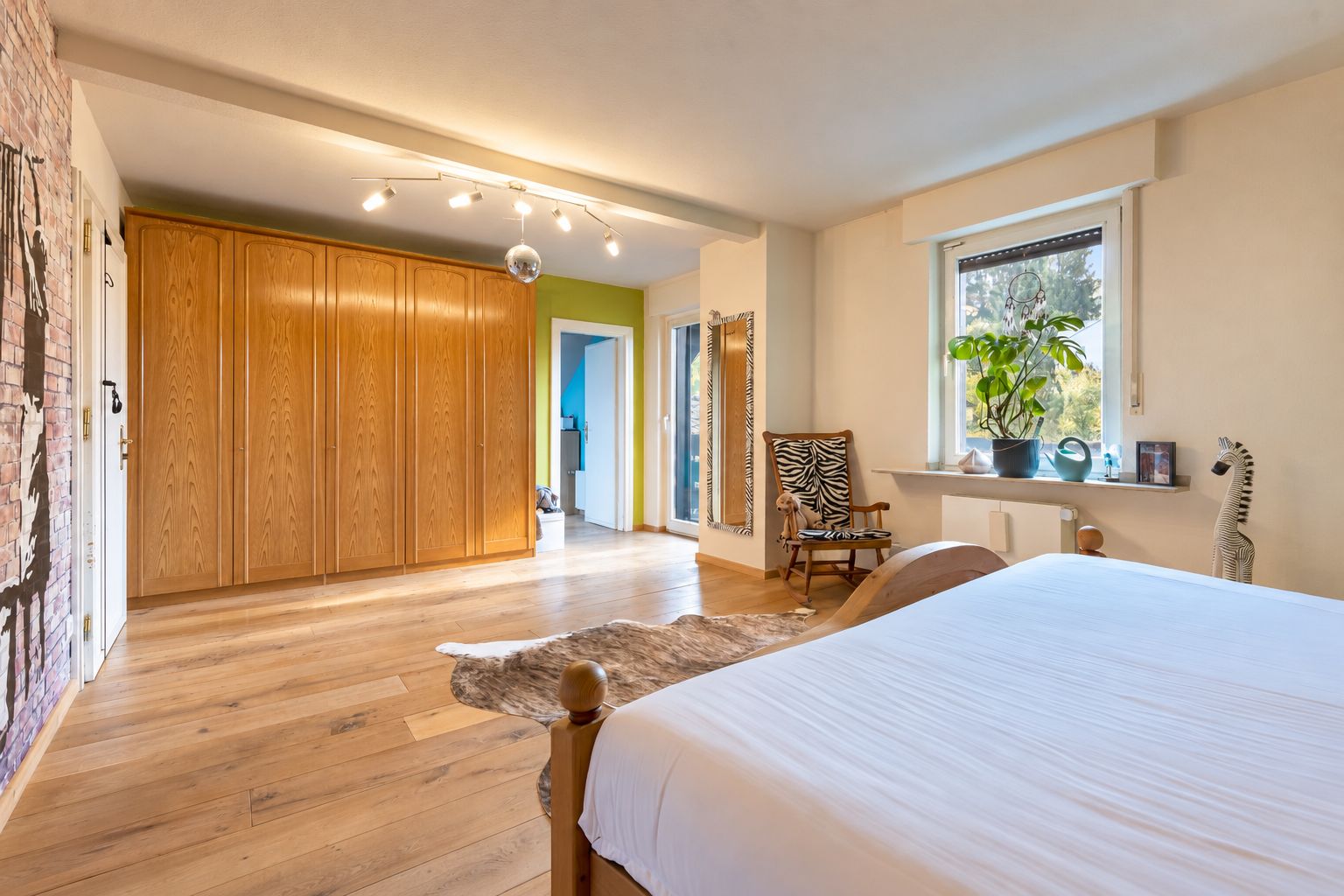 Helles Schlafzimmer mit Holzboden, großem Kleiderschrank, Bett und Fensterpflanze.
