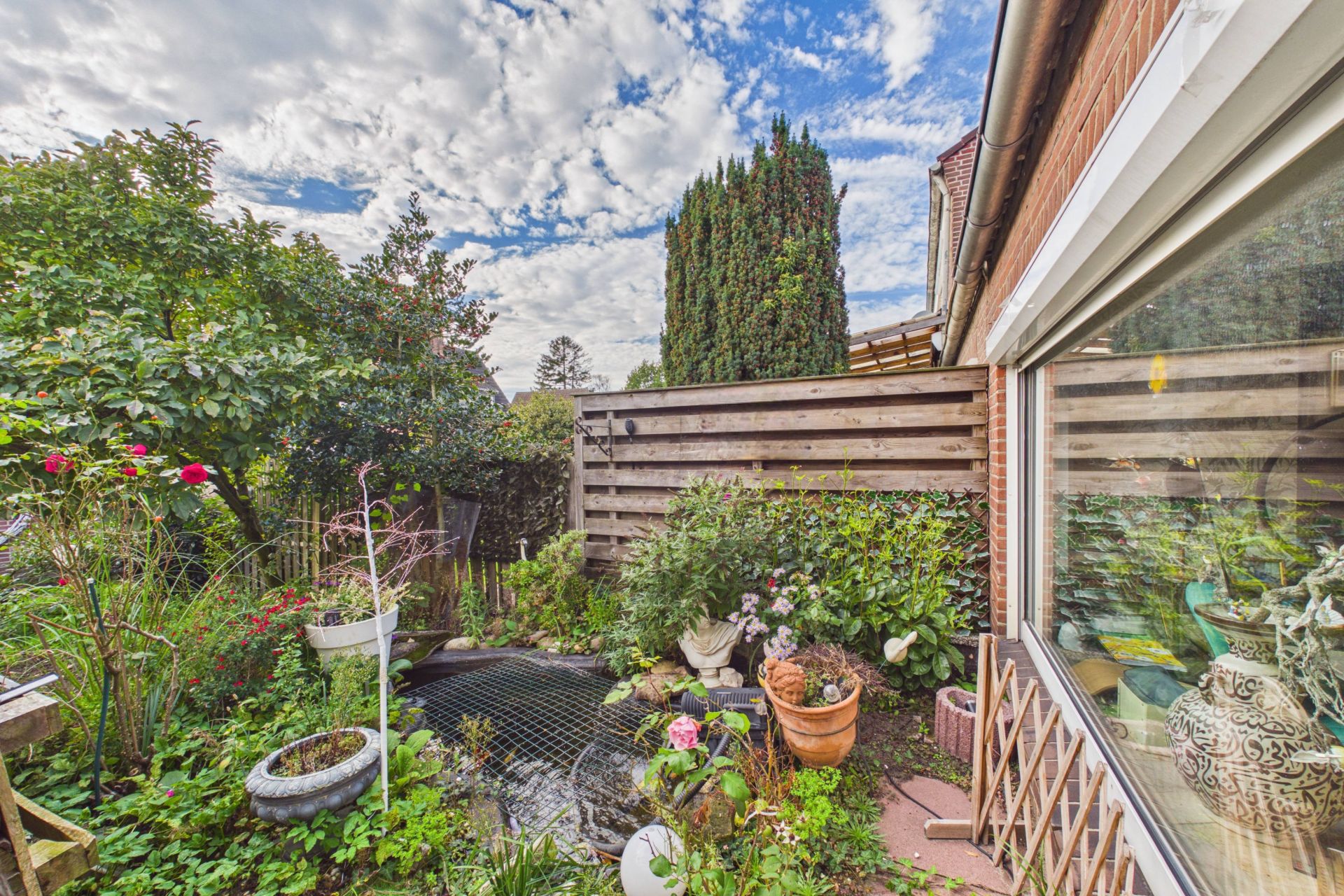 Kleiner Garten mit Teich, Blumen, Pflanzen und Holzlattenzaun neben einem Haus.