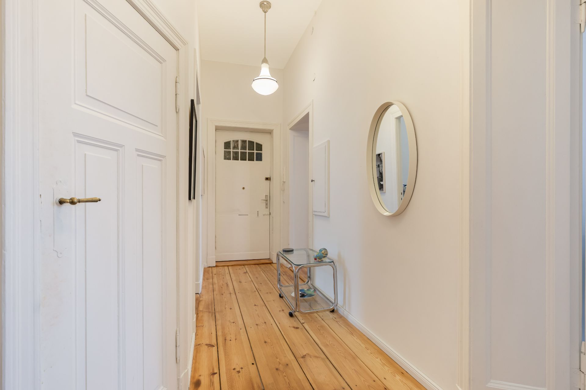 Heller Flur mit Holzboden, Wandspiegel, kleinem Tisch und weißer Eingangstür.