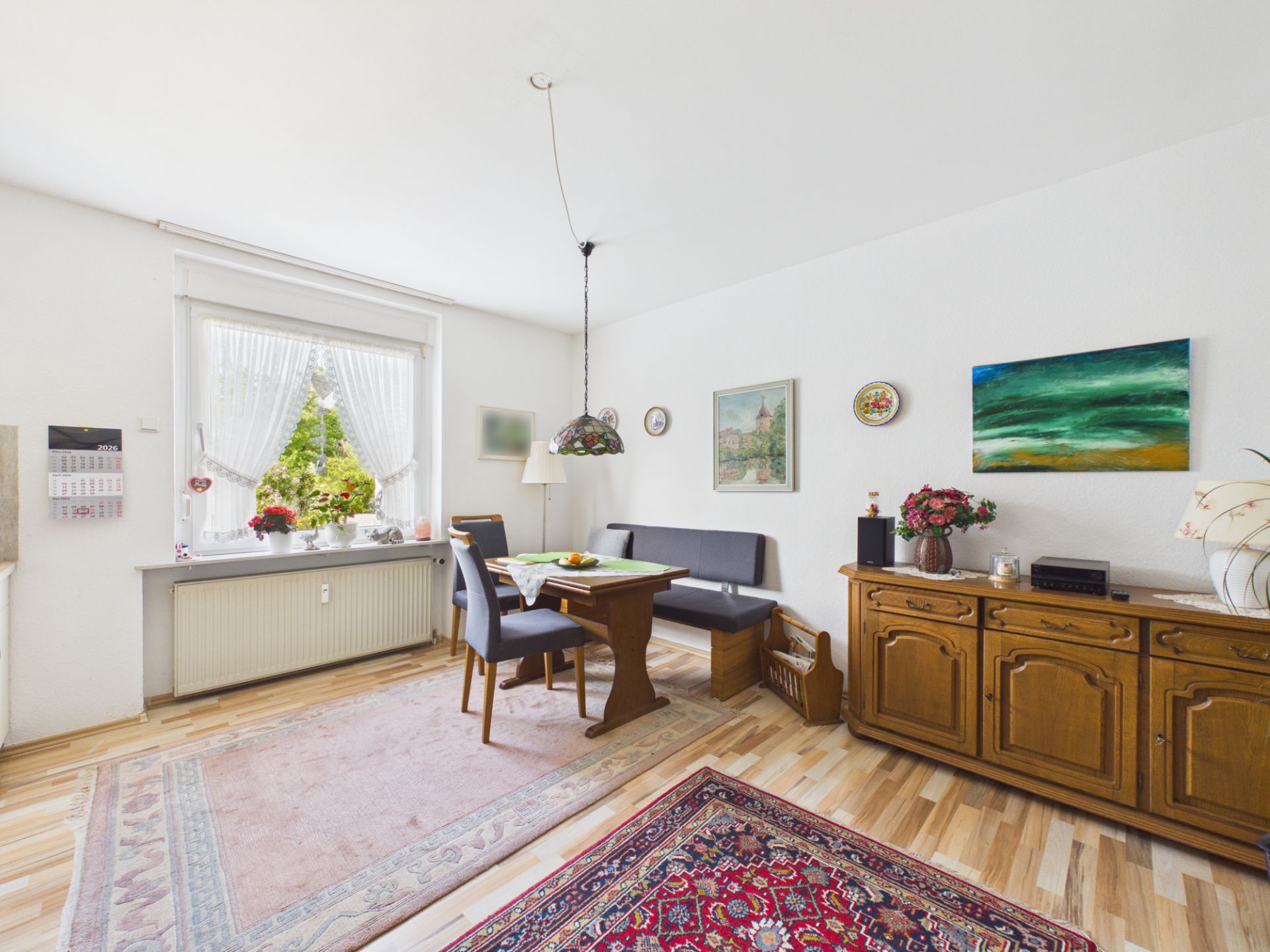 Helles Wohnzimmer mit Esstisch, Eckbank, Teppichen und Sideboard, Fenster mit Vorhängen.