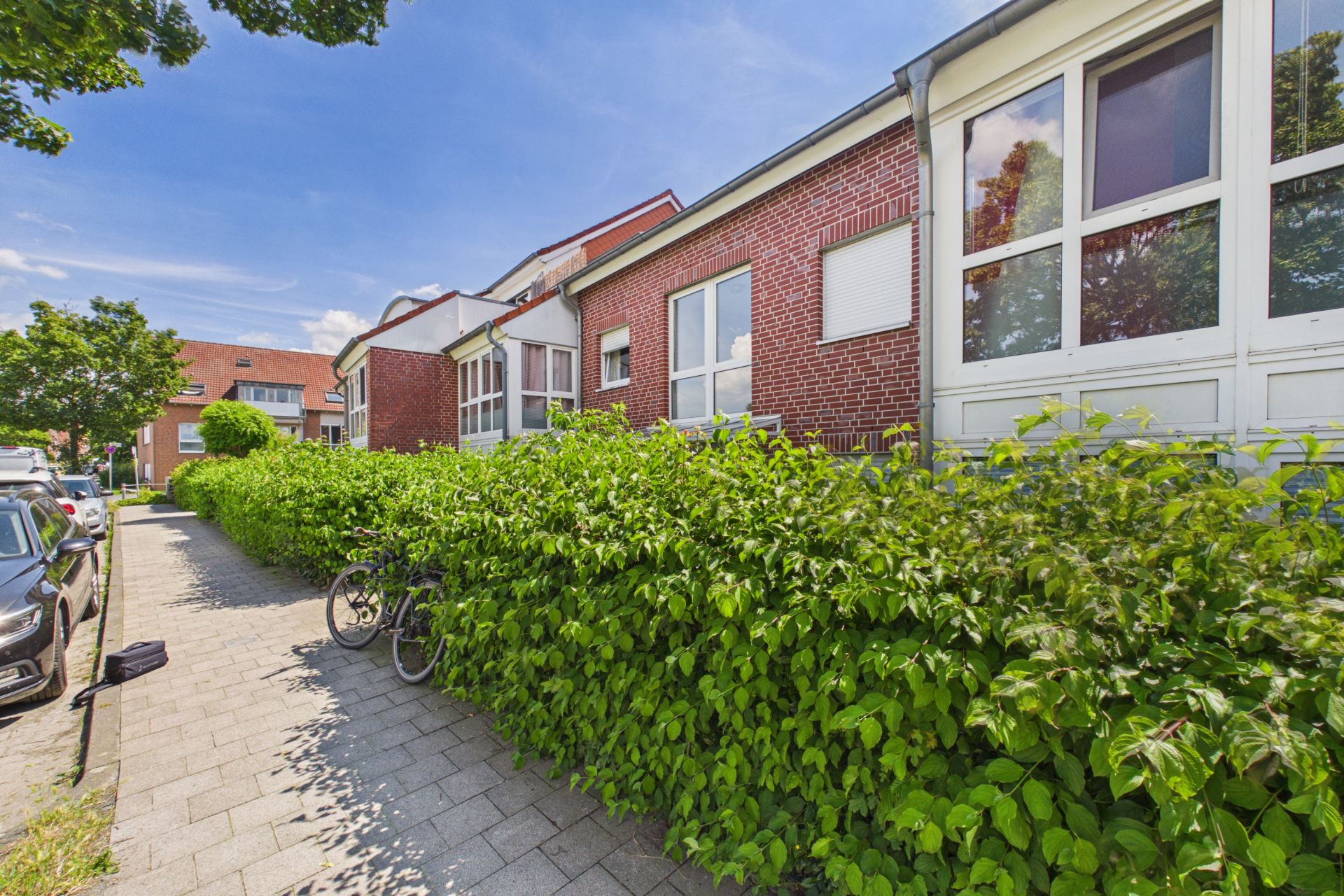 Backsteinwohnhaus mit großen Fenstern, Hecke und Fahrrad an einem Gehweg bei sonnigem Wetter.