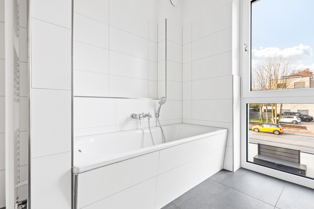 Helles Badezimmer mit weiß gefliester Badewanne und großem Fenster zur Straße.