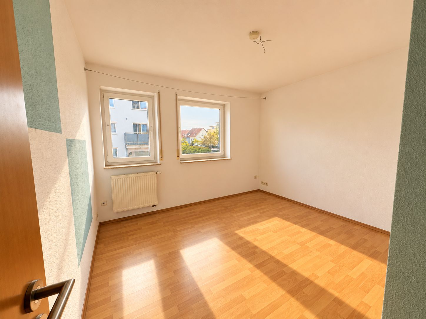 Helles, leeres Zimmer mit zwei Fenstern, Holzfußboden und weiß gestrichenen Wänden.