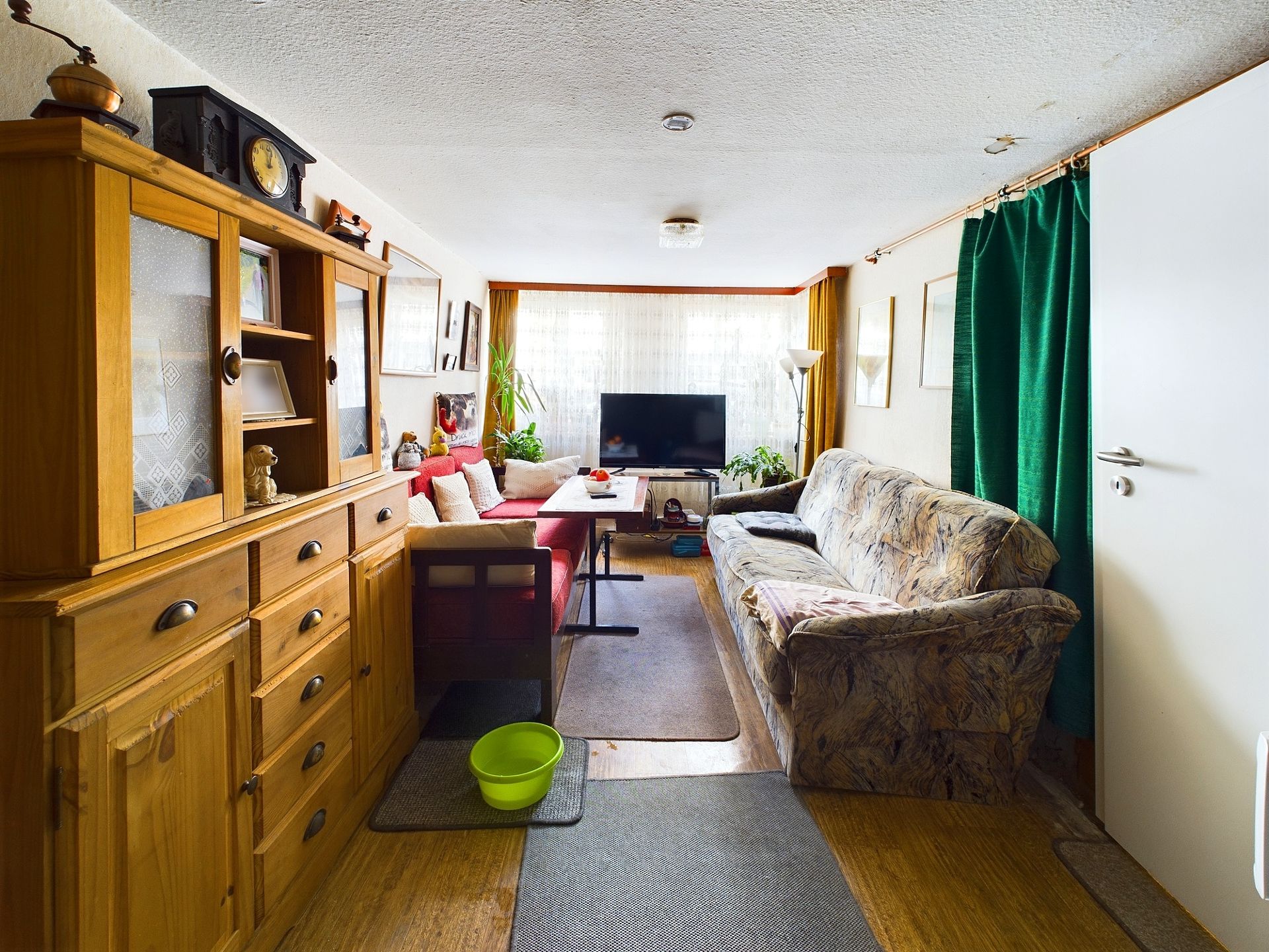 Wohnzimmer mit Sofa, Holzschrank, Fernseher und großem Fenster, grüne Gardine und Eimer auf dem Boden.