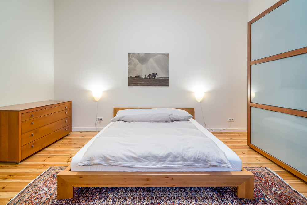 Schlafzimmer mit Holzbett, Kommode, Teppich, Wandbild und zwei Wandleuchten.
