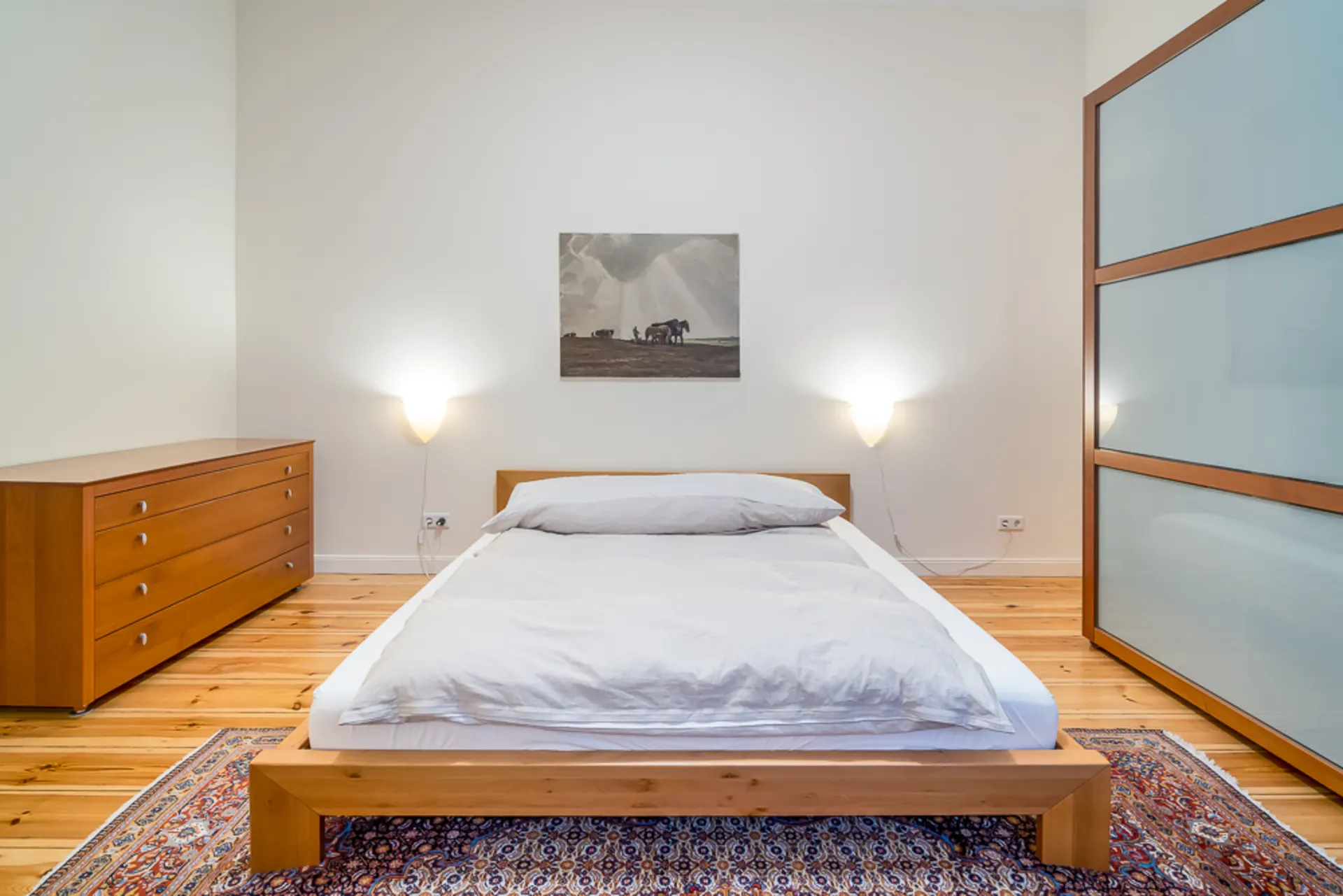 Schlafzimmer mit Holzbett, Kommode, Teppich, Wandbild und zwei Wandleuchten.