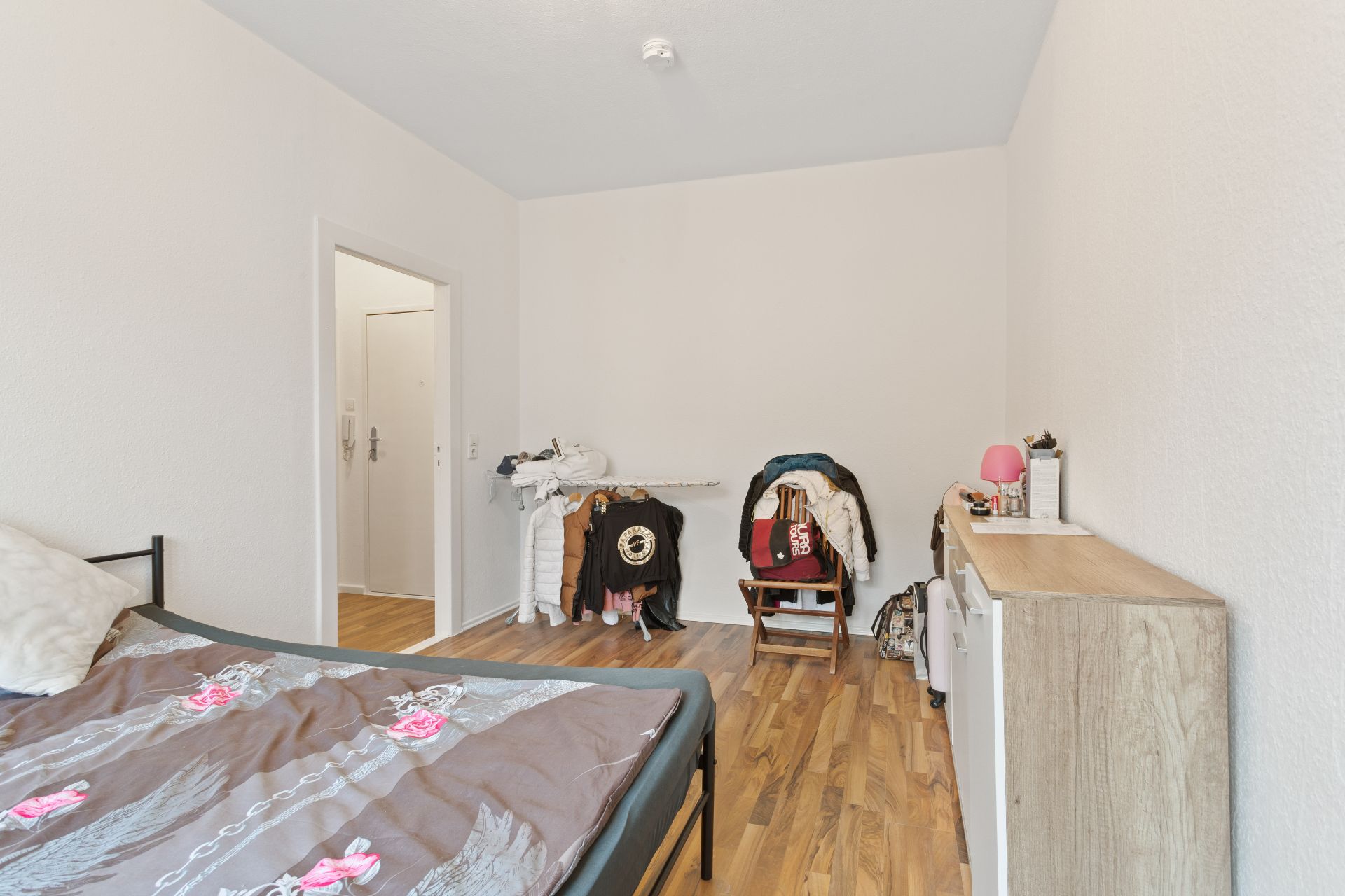 Schlafzimmer mit Bett, Kommode, Kleidungsstücken, Holzfußboden und offener Tür zum Flur.