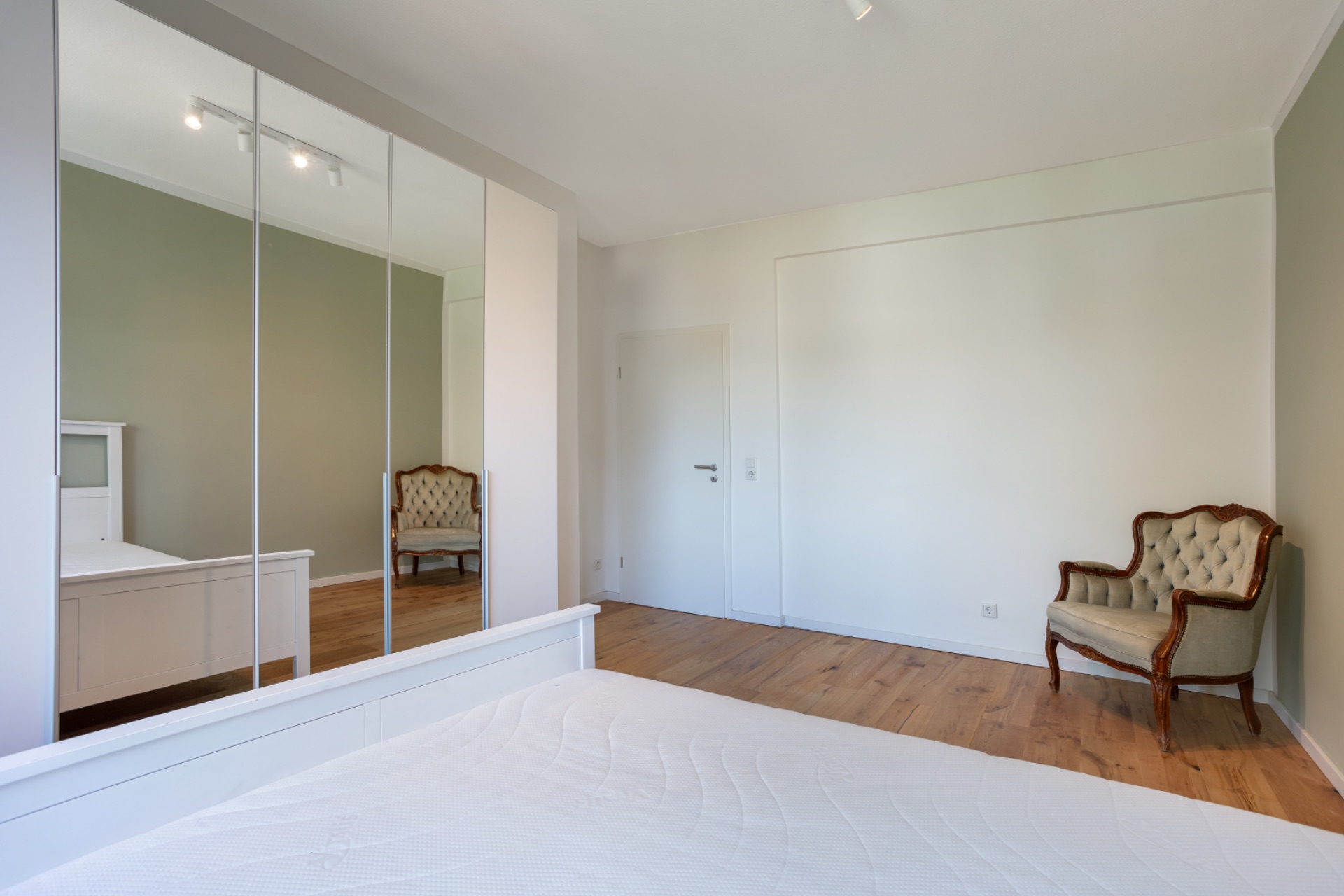 Helles Schlafzimmer mit Spiegelkleiderschrank, Bett und zwei Polsterstühlen auf Holzboden.