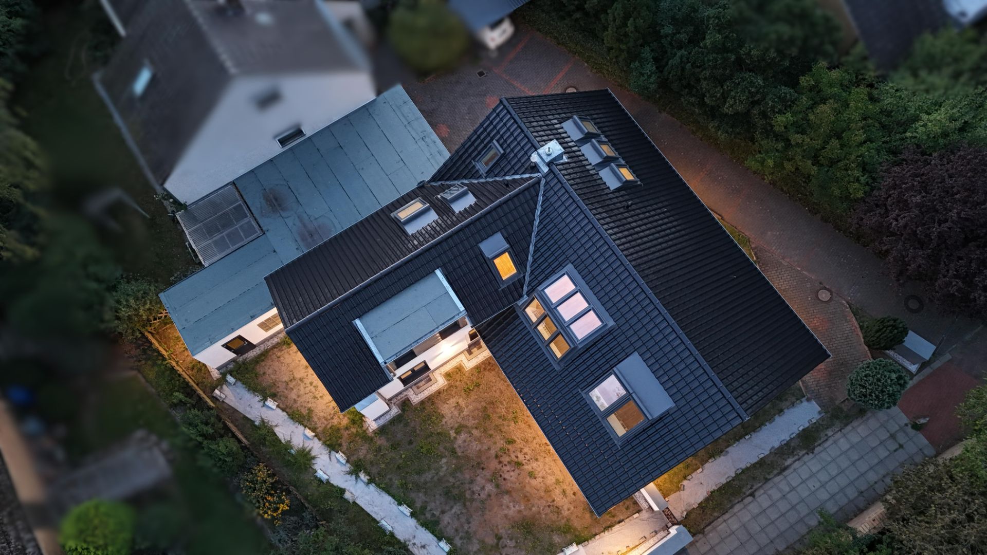 Draufsicht auf ein beleuchtetes Einfamilienhaus mit dunklem Dach und Gartenweg.