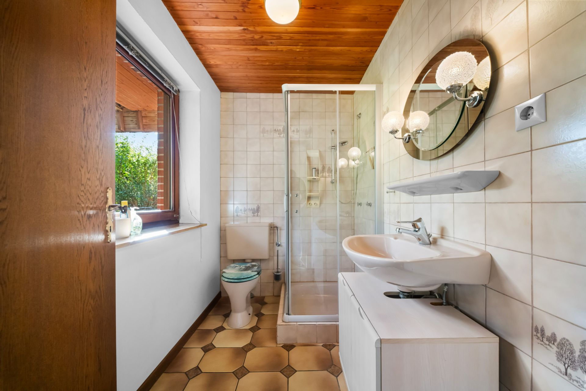Helles Badezimmer mit Dusche, Waschbecken, WC und Fenster. Holzbalkendecke und Fliesenboden.