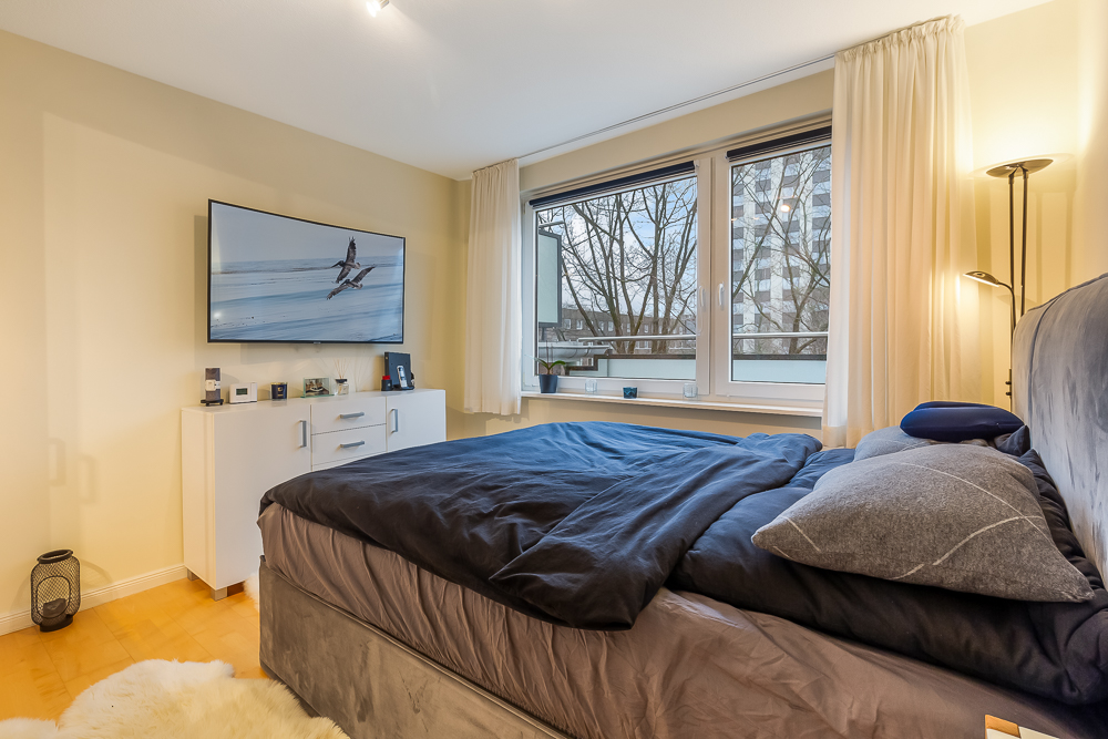 Schlafzimmer mit großem Bett, Kommode, Fernseher und zwei großen Fenstern mit Vorhängen.