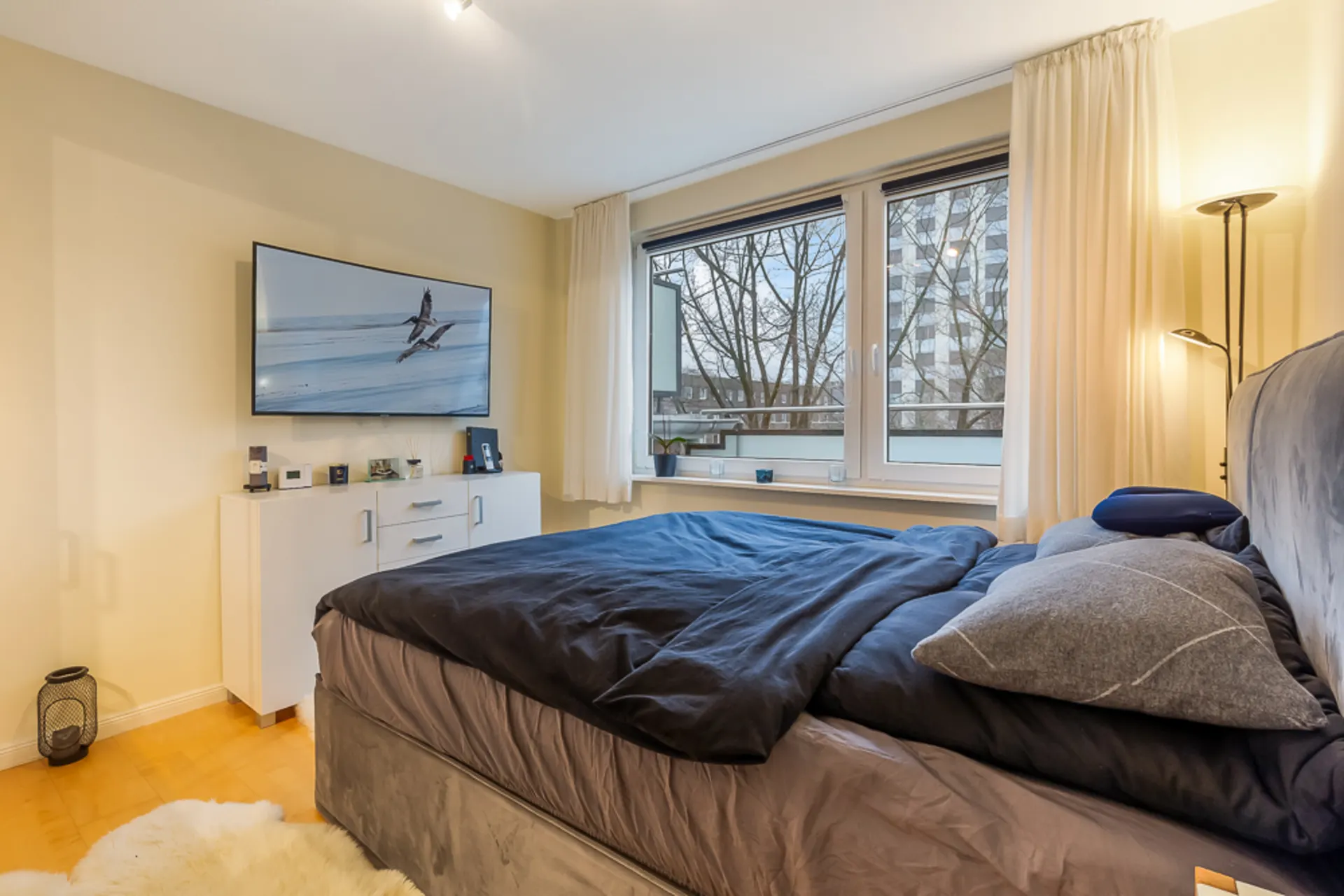Schlafzimmer mit großem Bett, Kommode, Fernseher und zwei großen Fenstern mit Vorhängen.