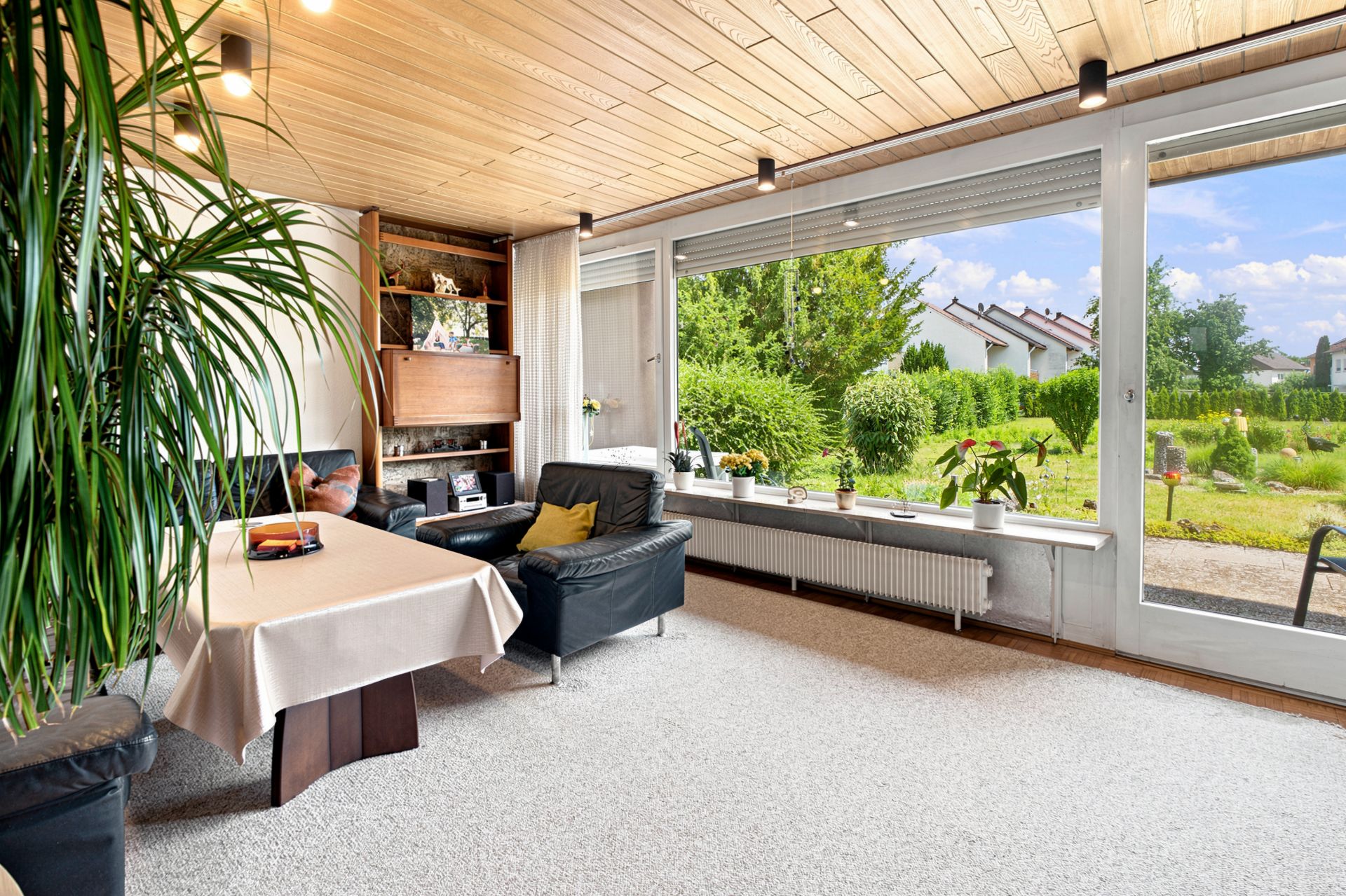 Helles Wohnzimmer mit großen Fenstern, schwarzen Sesseln, Tisch und Blick in den grünen Garten.