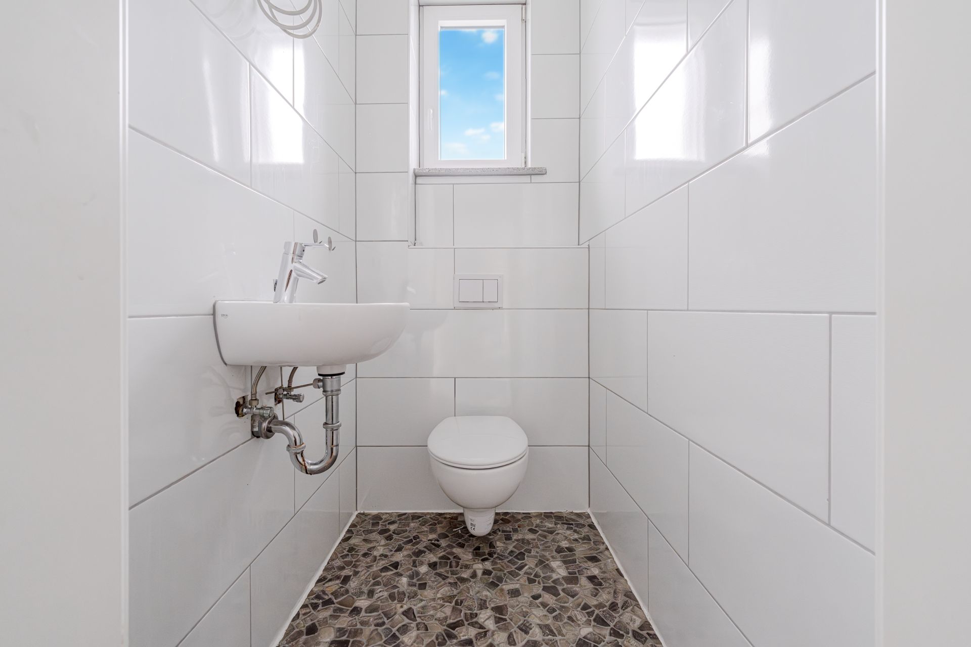 Kleines Badezimmer mit weiß gefliesten Wänden, WC, Waschbecken und kleinem Fenster.