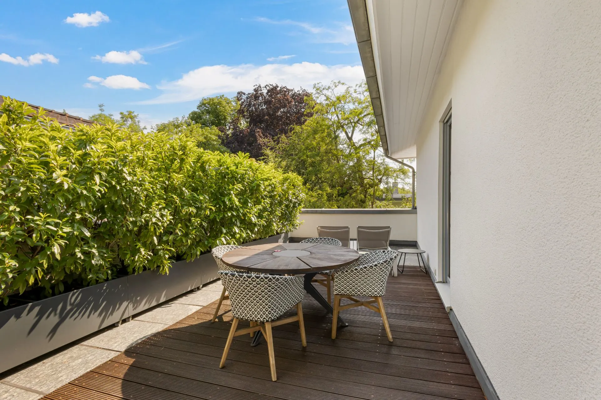 Große Terrasse mit Holzboden, rundem Tisch und Stühlen, umgeben von grünen Pflanzen.