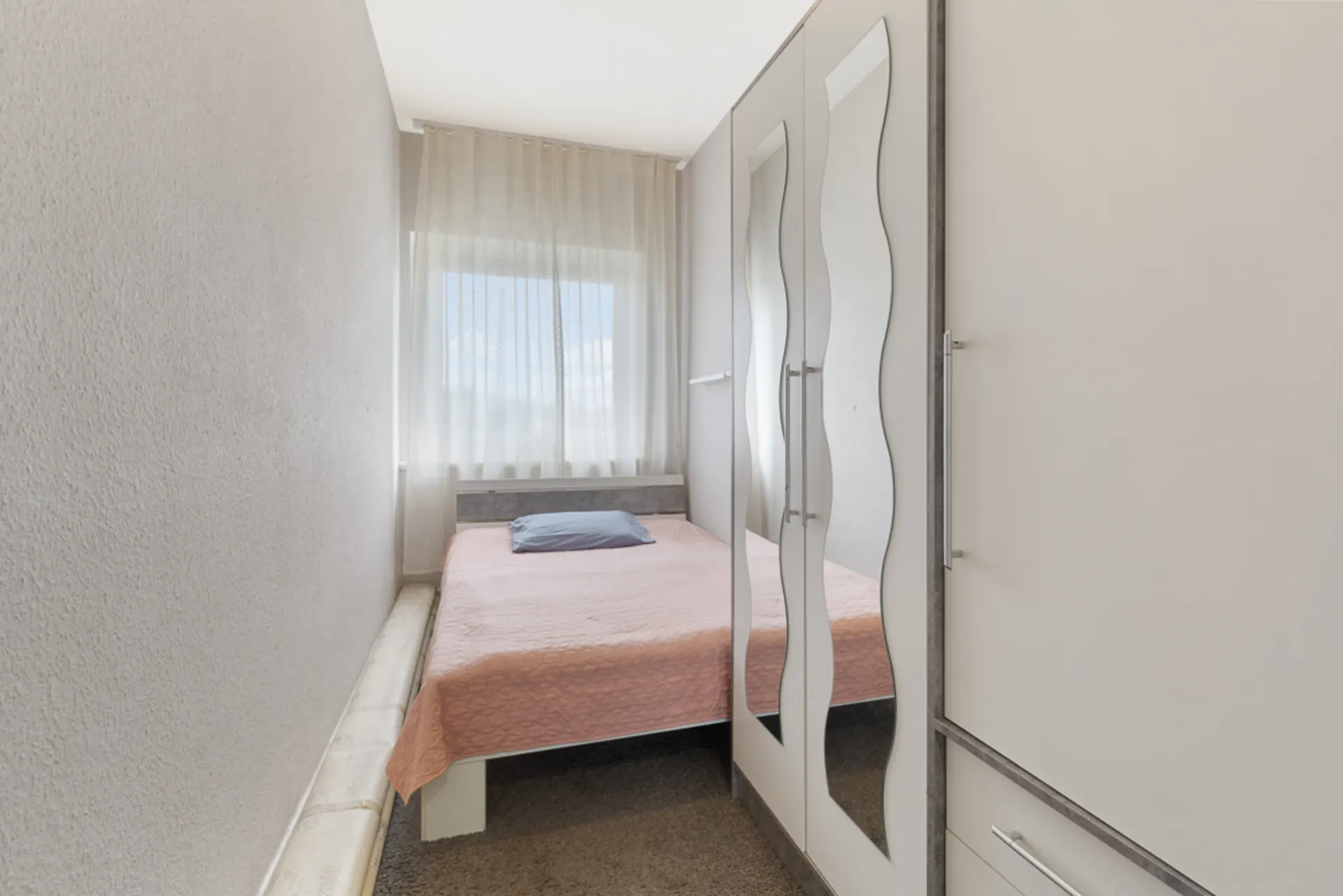 Schmales Schlafzimmer mit Bett, Spiegelschrank und Fenster mit hellem Vorhang.
