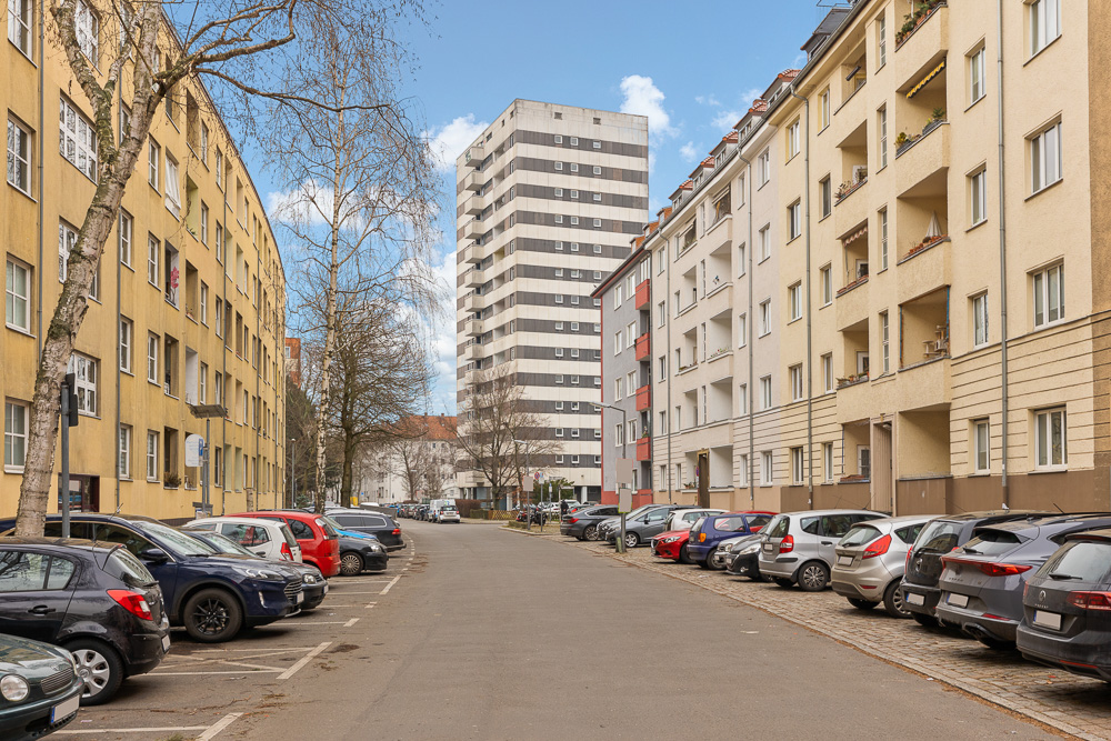 Straße mit parkenden Autos, mehrstöckigen Wohnhäusern und Bäumen auf beiden Seiten.