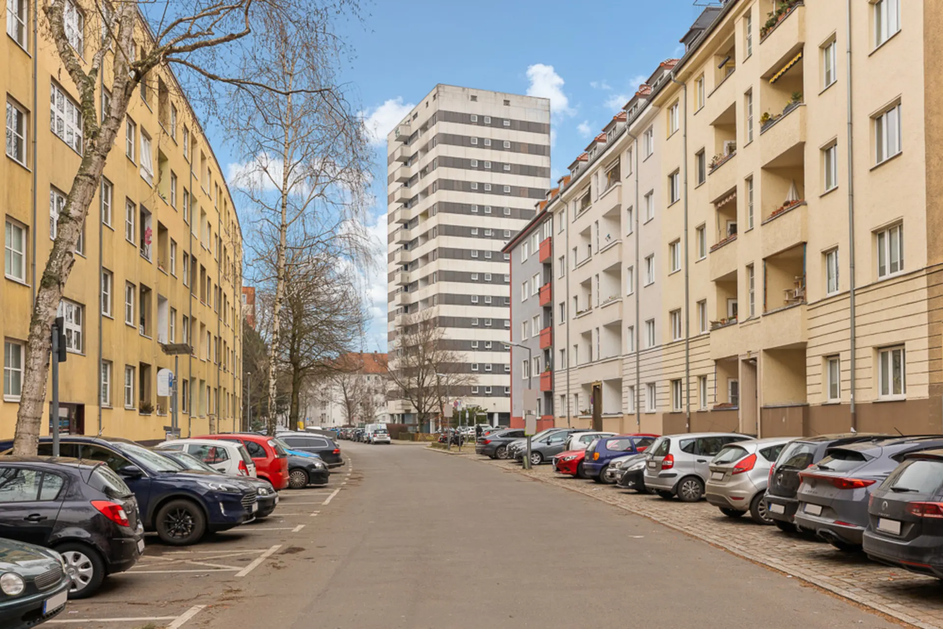 Straße mit parkenden Autos, mehrstöckigen Wohnhäusern und Bäumen auf beiden Seiten.