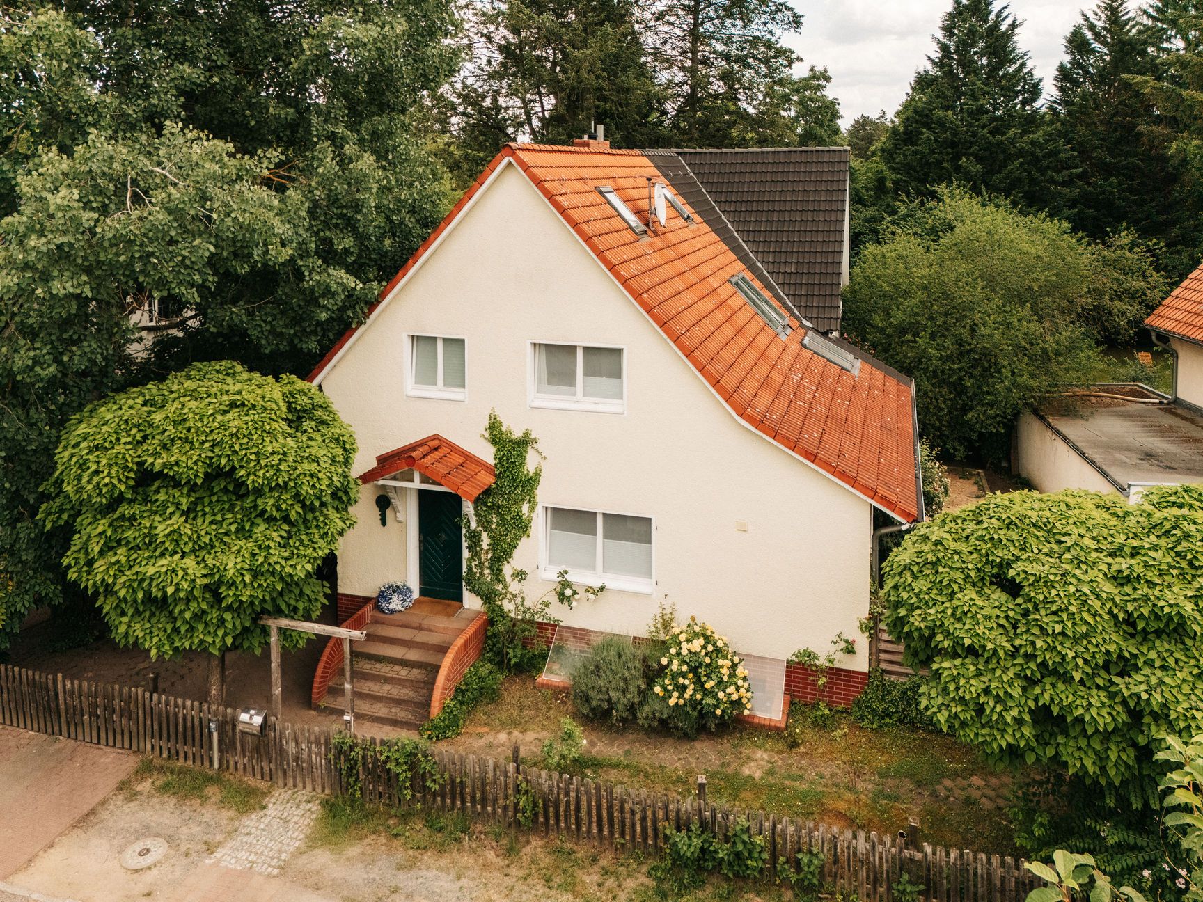 Einfamilienhaus mit rotem Dach und kleinem Garten, umgeben von Bäumen und Holzzaun.