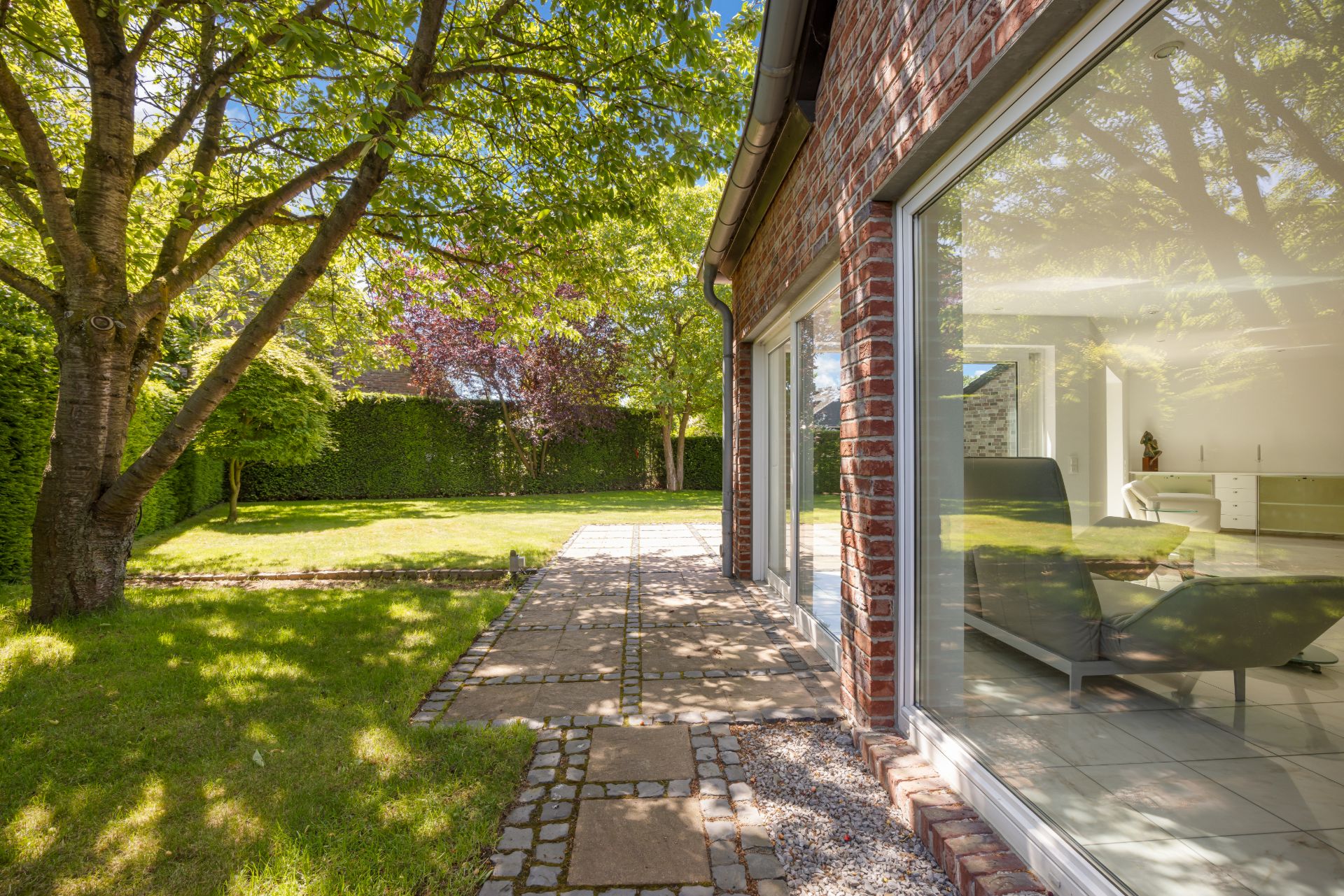 Garten mit Rasen, Bäumen und gepflastertem Weg neben einem Haus mit großen Fenstern.