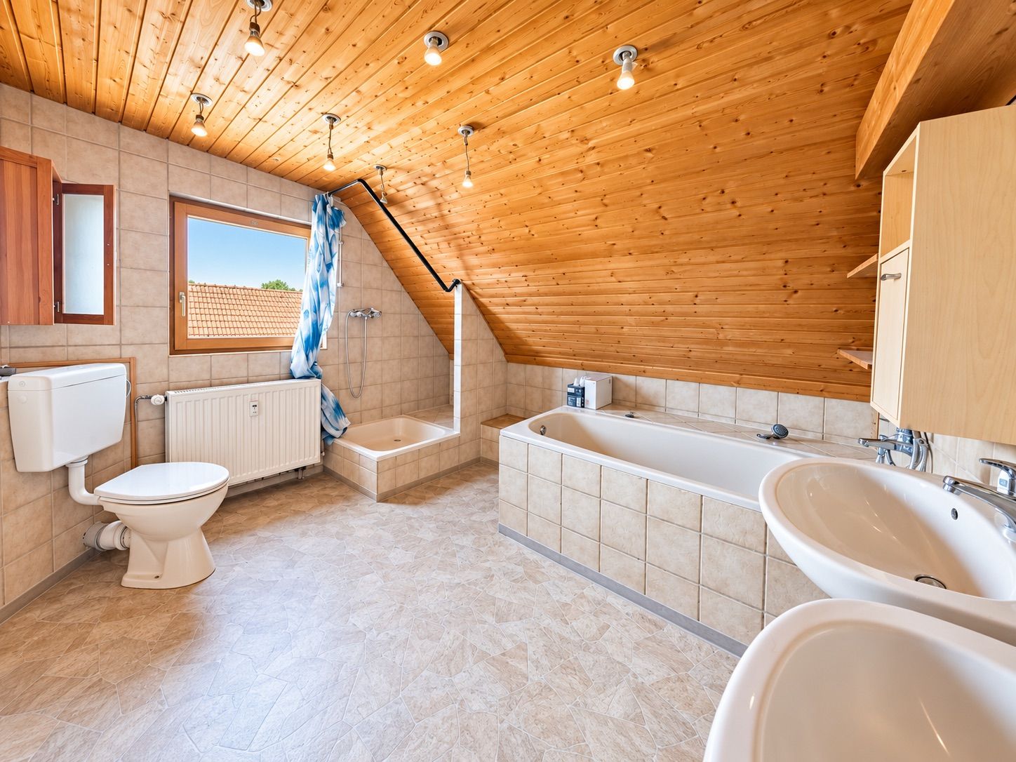 Badezimmer mit Dachschräge, Badewanne, Dusche, Toilette, zwei Waschbecken und Fenster.