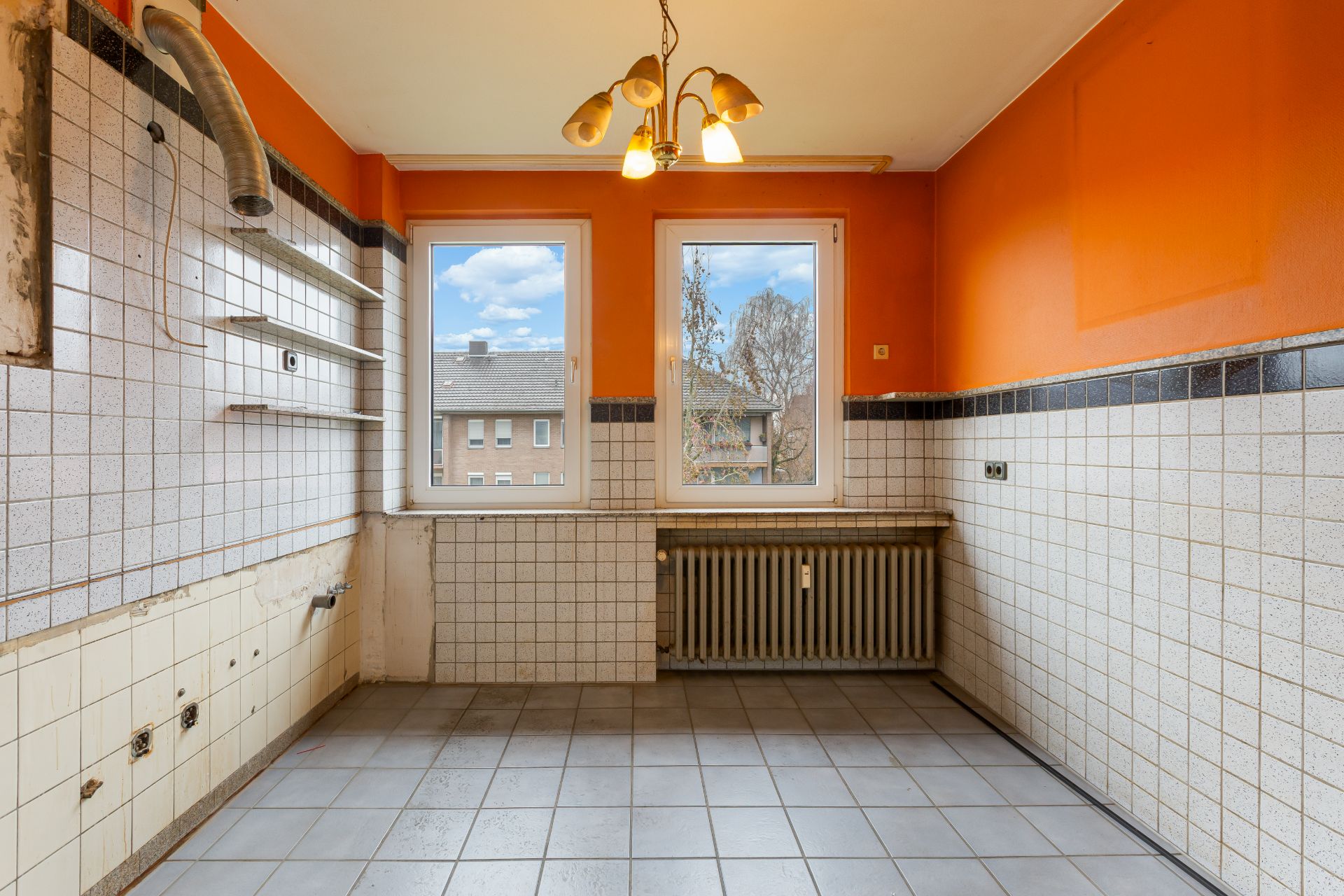 Leere Küche mit orangefarbenen Wänden, Fliesen, zwei Fenstern und Heizkörper.