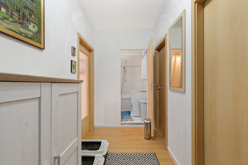 Heller Flur mit Holzfußboden, Spiegel, Türen und Blick ins Badezimmer mit Dusche.