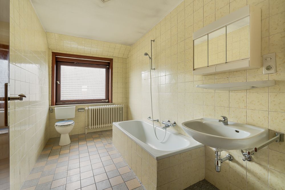 Helles Bad mit Badewanne, Waschbecken, Toilette und Fenster, Fliesen an Wand und Boden.