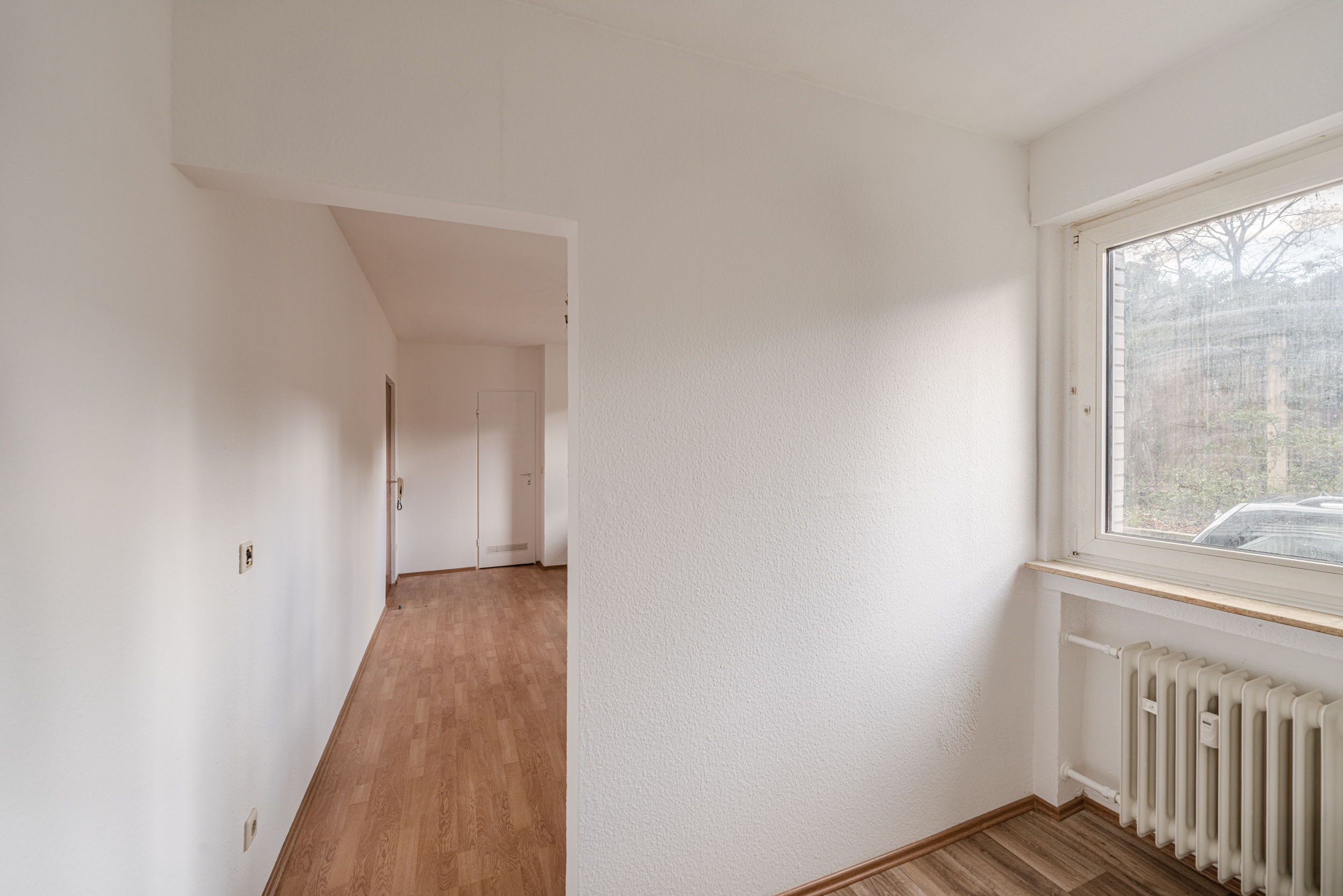 Heller Flur mit Holzboden, Fenster und Heizkörper, Blick auf eine weiße Tür.