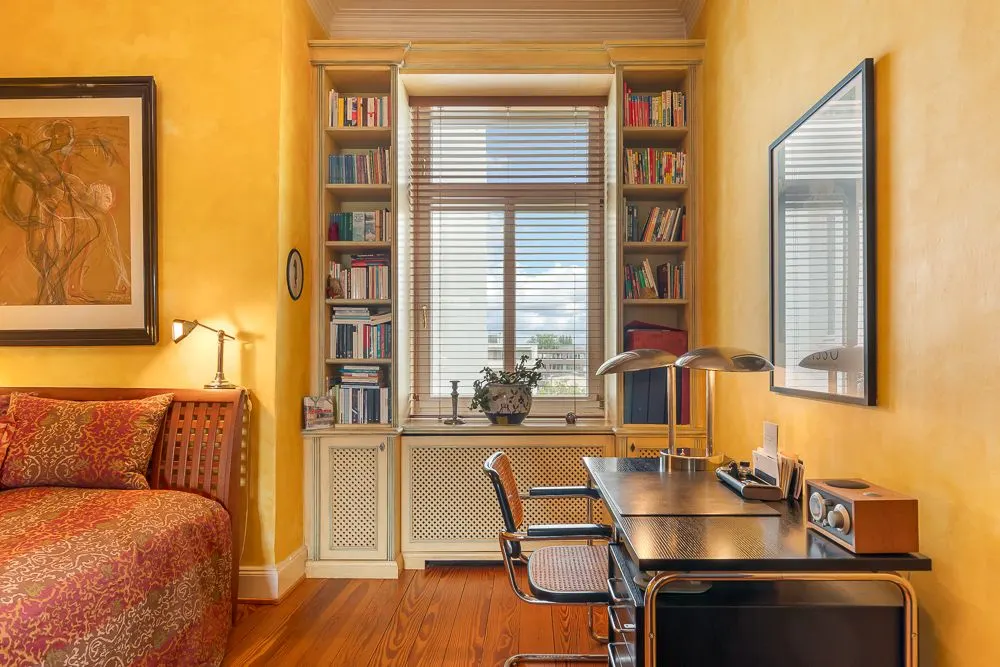 Arbeitszimmer mit Schreibtisch, Stuhl, Regal voller Bücher und Fenster mit Jalousien.