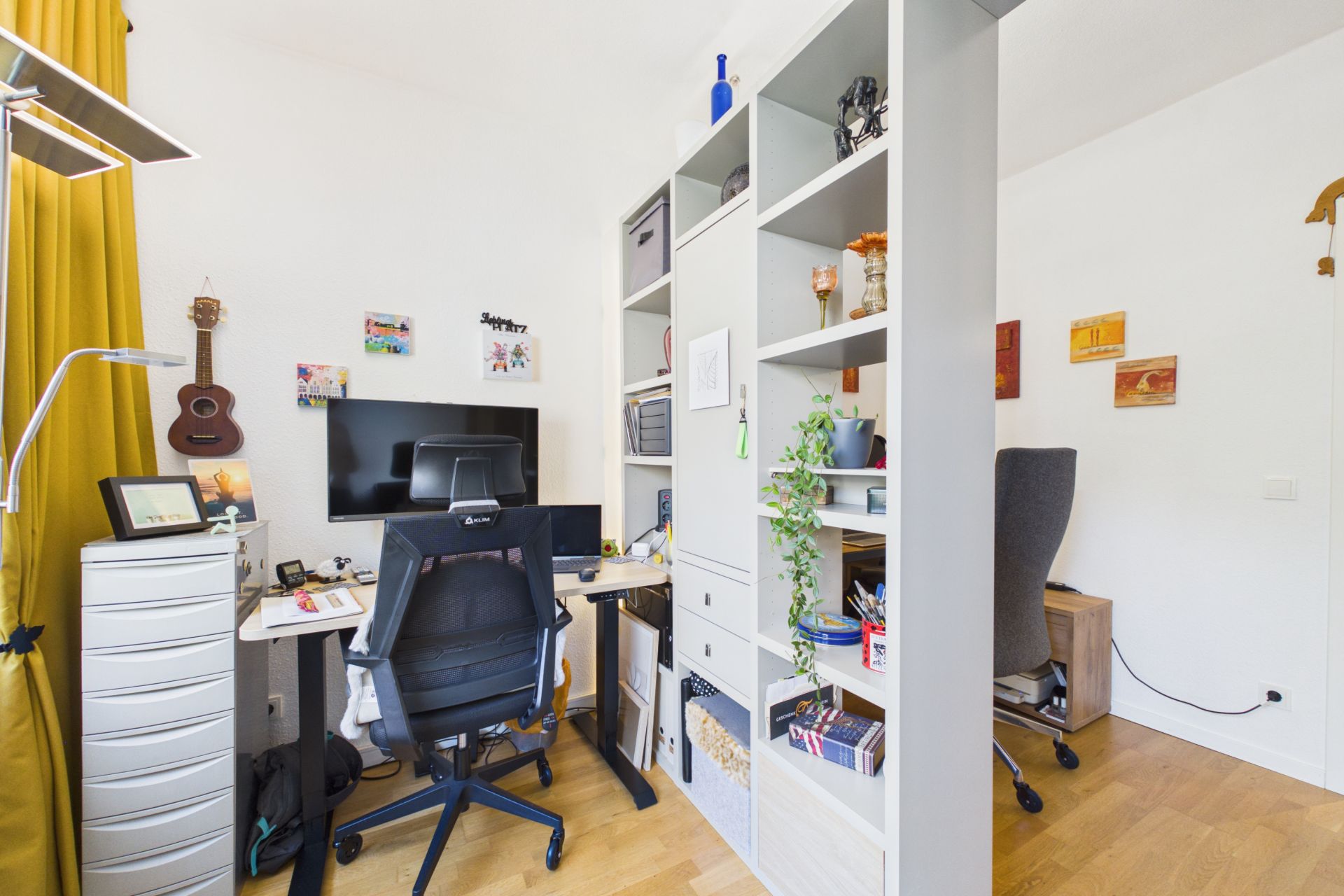 Helles Arbeitszimmer mit Schreibtisch, Stuhl, Regal und Computer, dekoriert mit Pflanzen und Bildern.