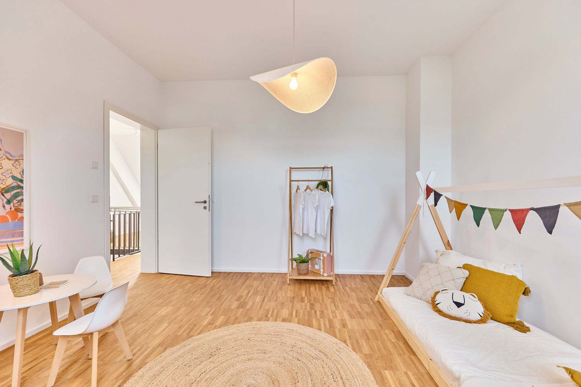 Helles Kinderzimmer mit Holzboden, niedriger Bettcouch, Kleiderständer und kleinem Tisch mit zwei Stühlen.