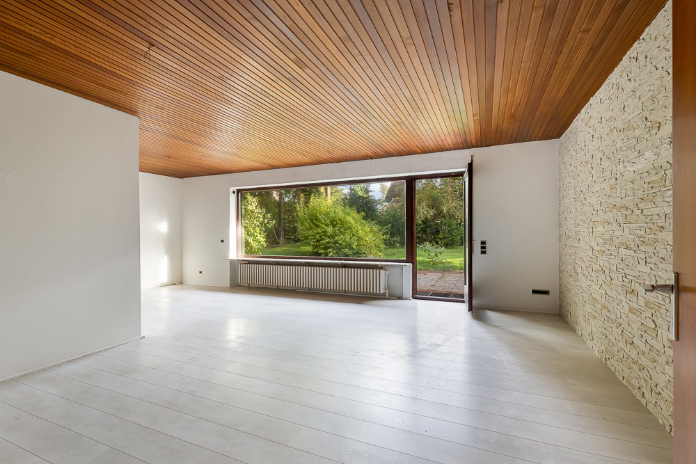 Helles, leeres Wohnzimmer mit Holzdecke, weißem Boden und großem Fenster mit Blick in den Garten.