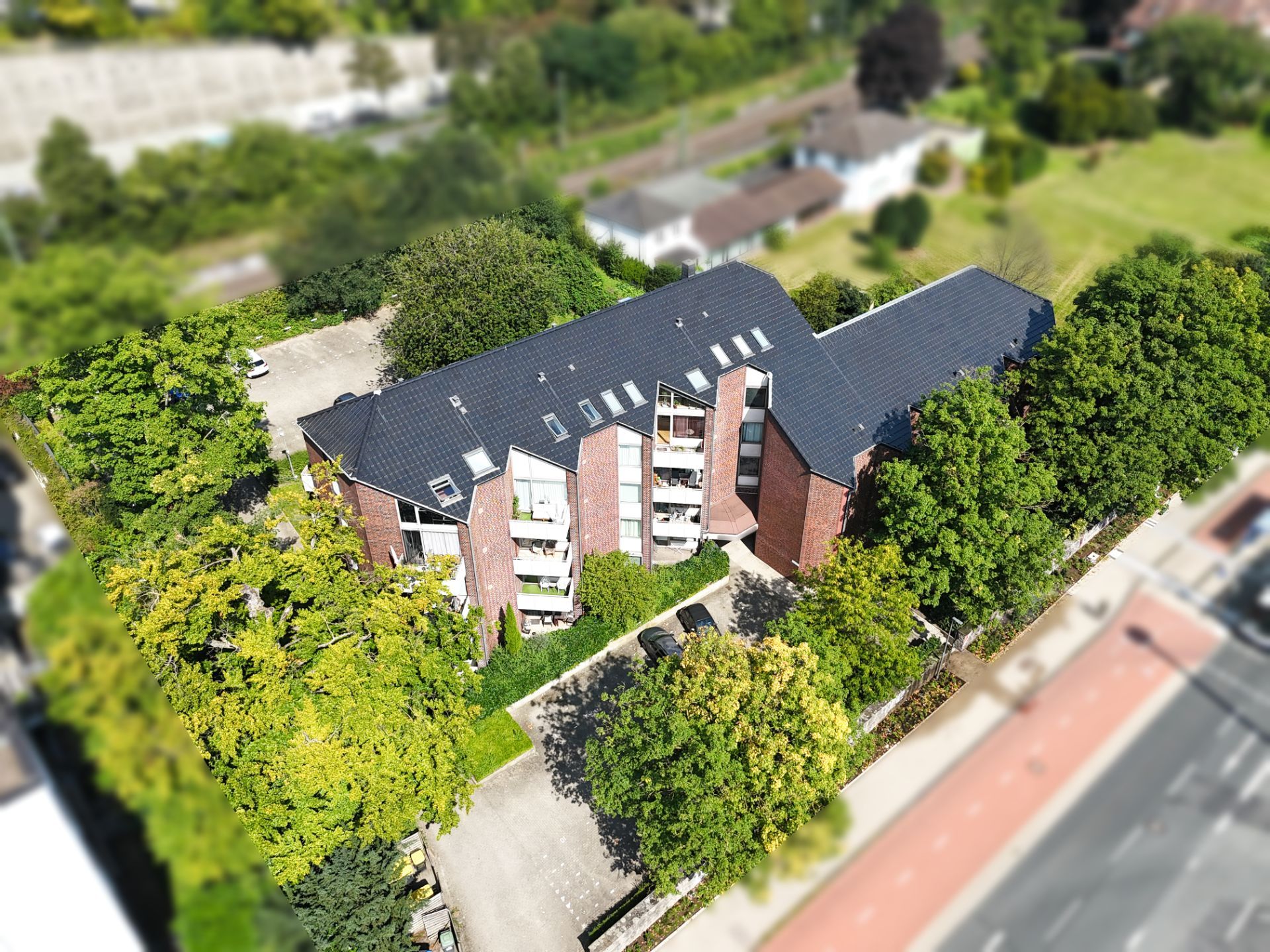 Mehrfamilienhaus mit dunklem Dach, umgeben von Bäumen und Parkplätzen, aus der Vogelperspektive.