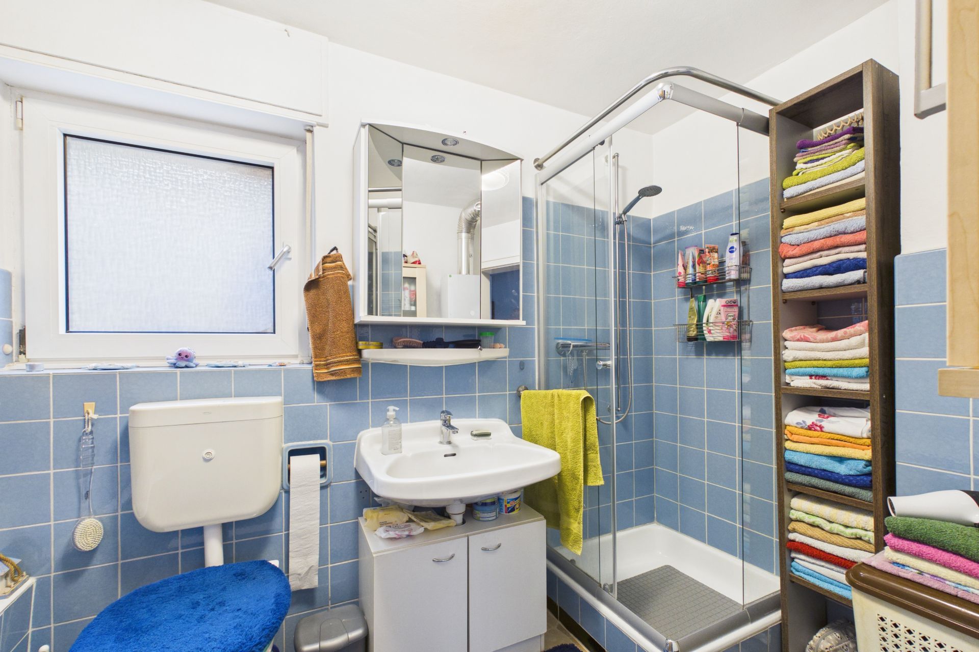 Kleines Badezimmer mit Dusche, Waschbecken, Toilette und Regal mit bunten Handtüchern.