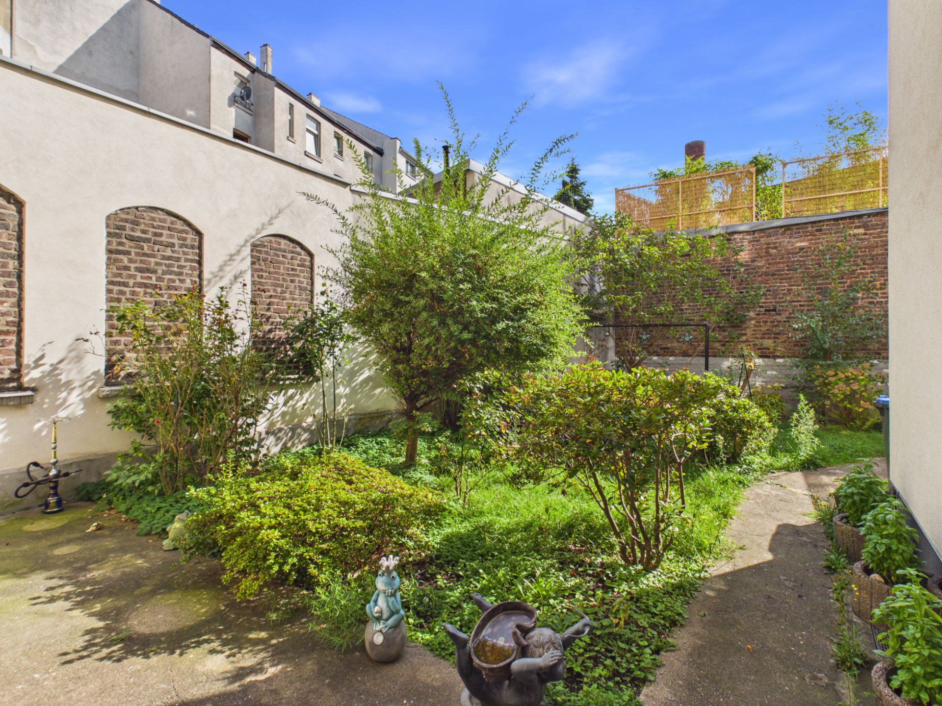Kleiner Innenhof mit Pflanzen, Büschen, einer Figur und einem Gartenweg neben Hauswänden.