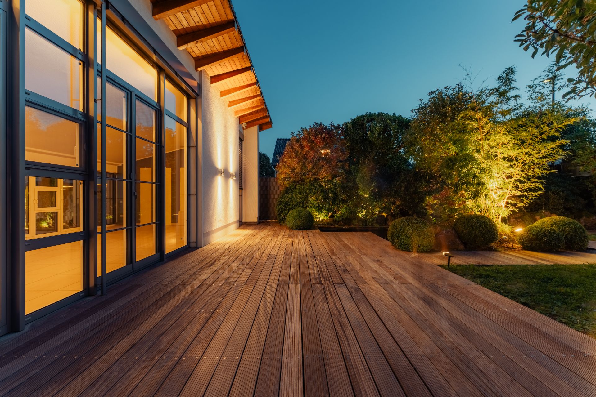 Große Holzterrasse mit Gartenbeleuchtung und bodentiefen Fenstern am Abend.