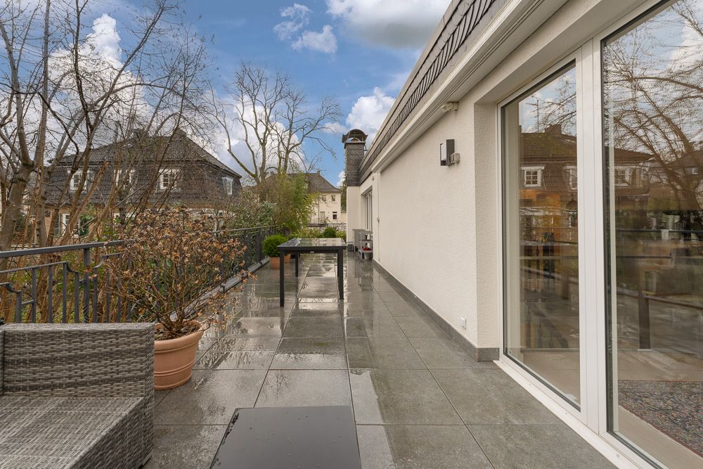 Große Terrasse mit Sitzbank, Tisch, Pflanzenkübel und Glasgeländer an einem Wohnhaus.