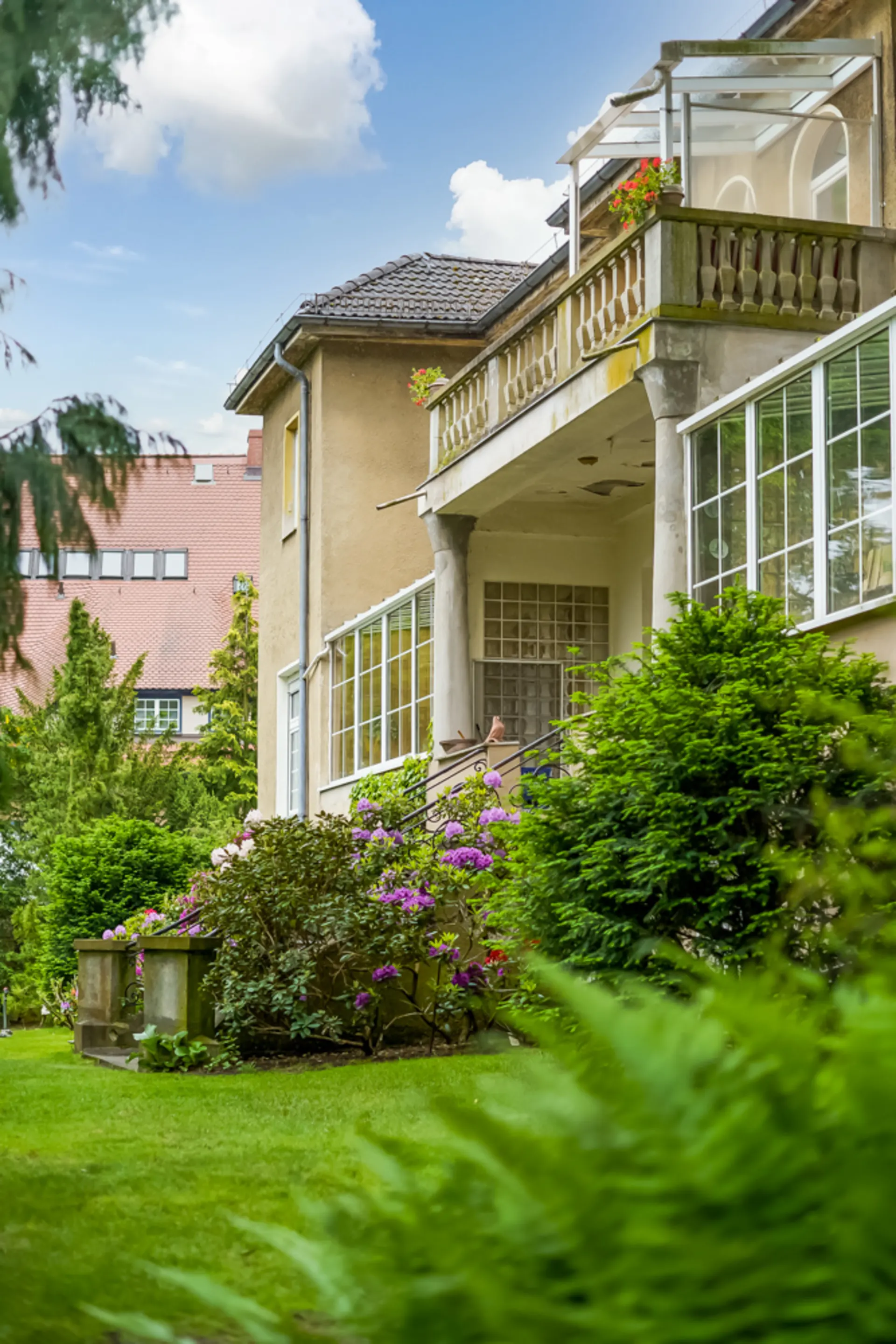 Wohnhaus mit Balkon, großen Fenstern und gepflegtem Garten mit blühenden Sträuchern.