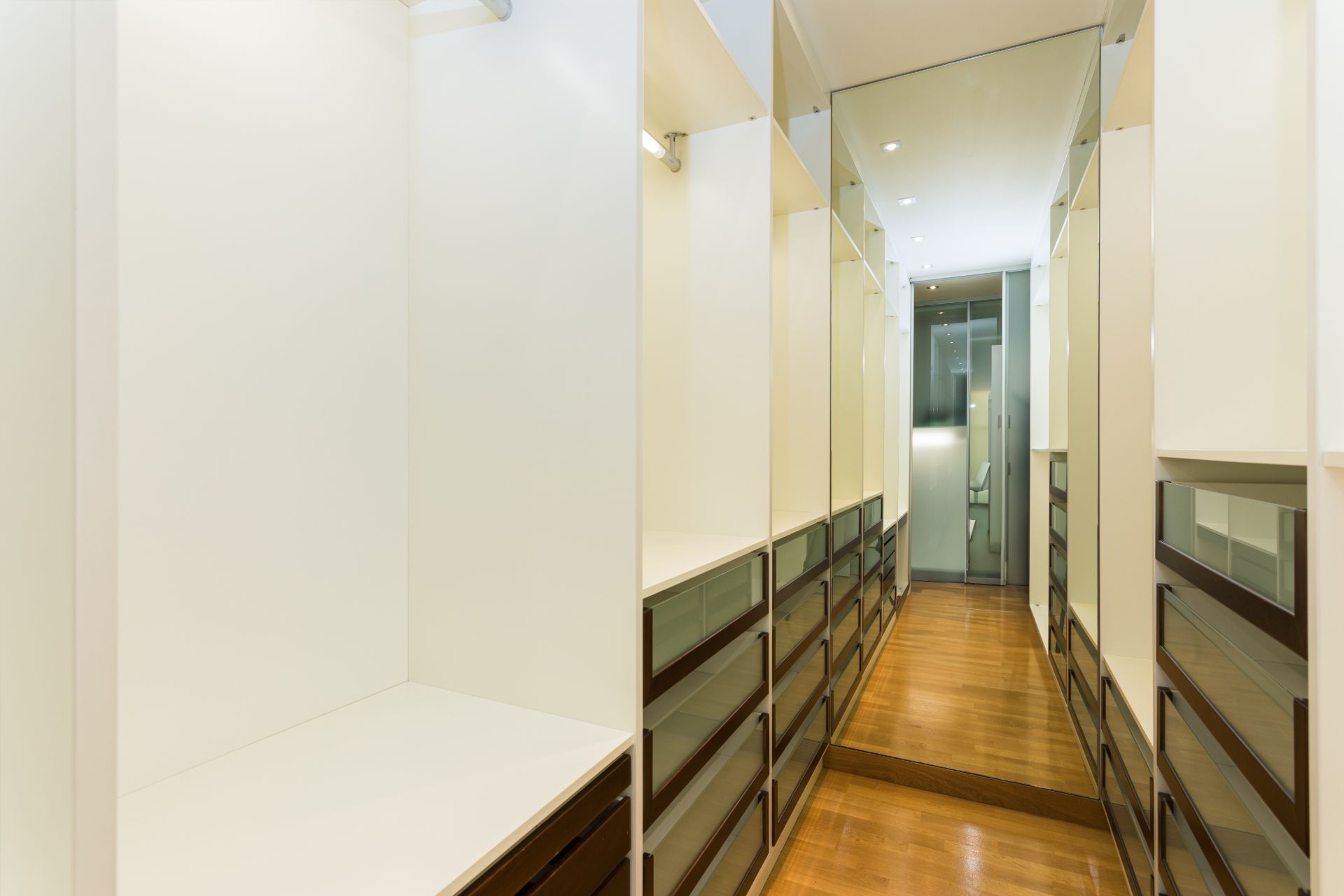 Moderner begehbarer Kleiderschrank mit weißen Regalen, Schubladen und einem großen Spiegel am Ende.