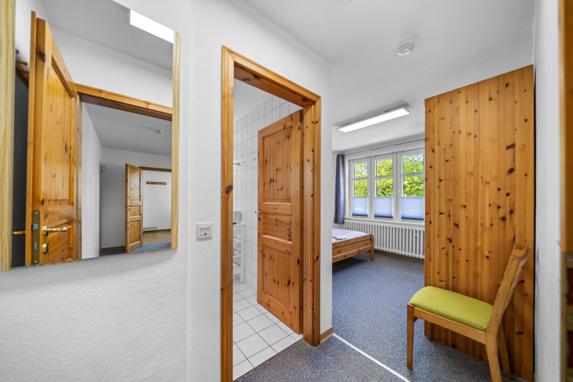 Heller Flur mit Holzinnentüren, Spiegel, Stuhl und Blick in ein Schlafzimmer mit Fenster.