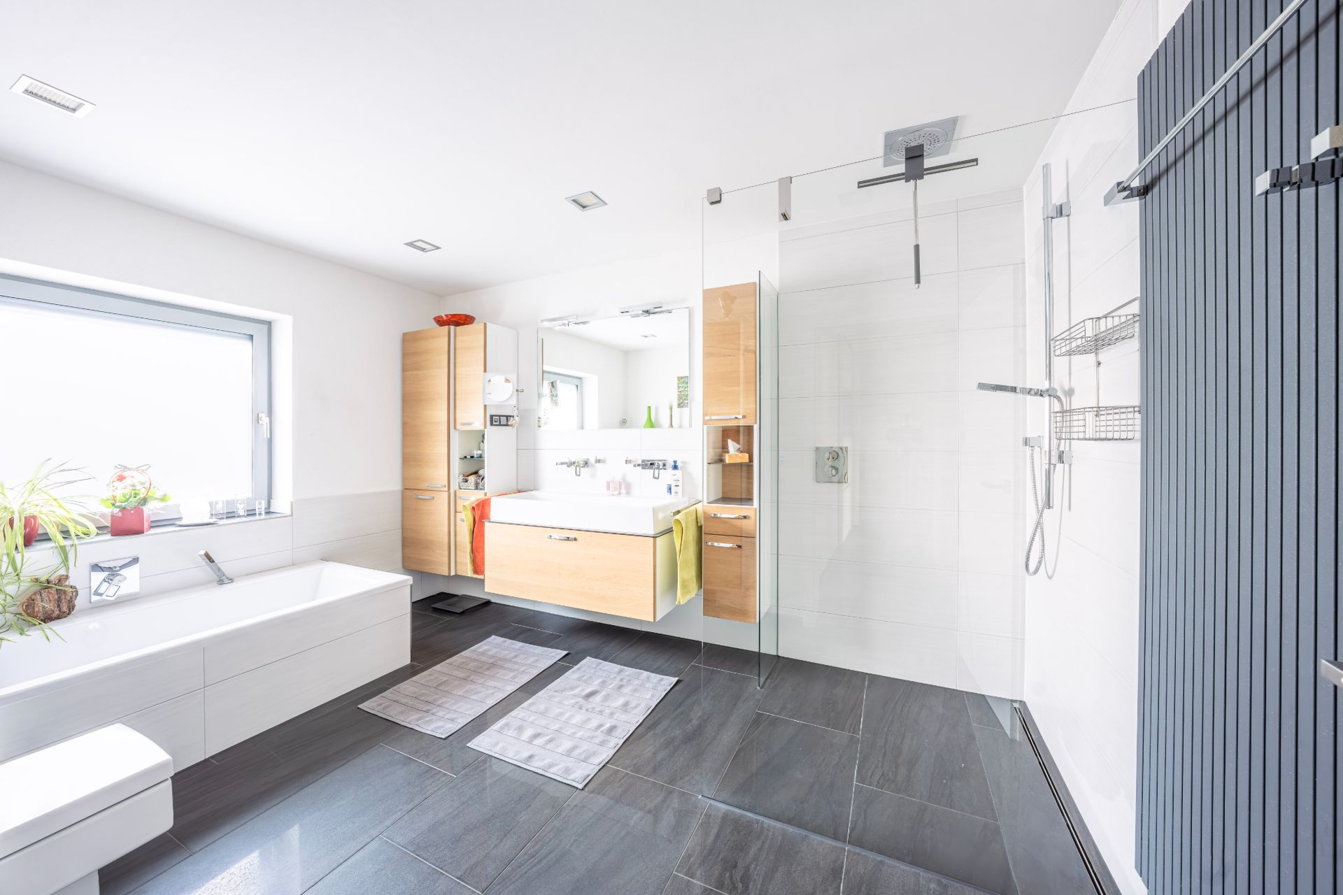 Helles Bad mit Badewanne, großer Dusche, Waschbecken und Fenster, schwarze Fliesen am Boden.