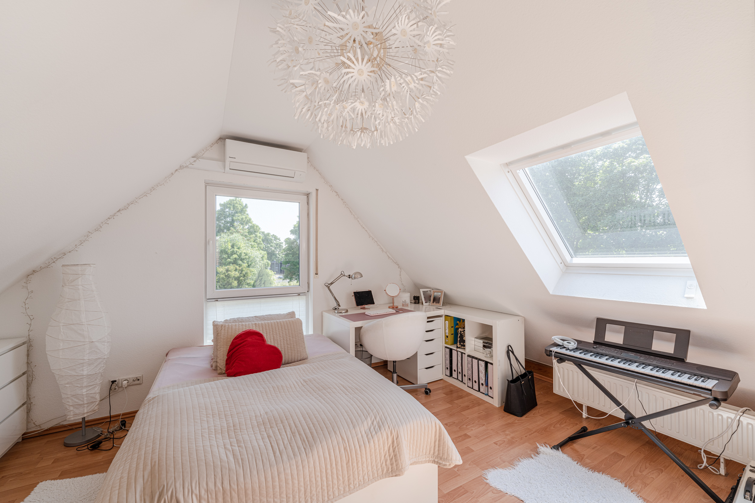 Helles Schlafzimmer mit Dachschräge, Bett, Schreibtisch, Fenster und E-Piano.