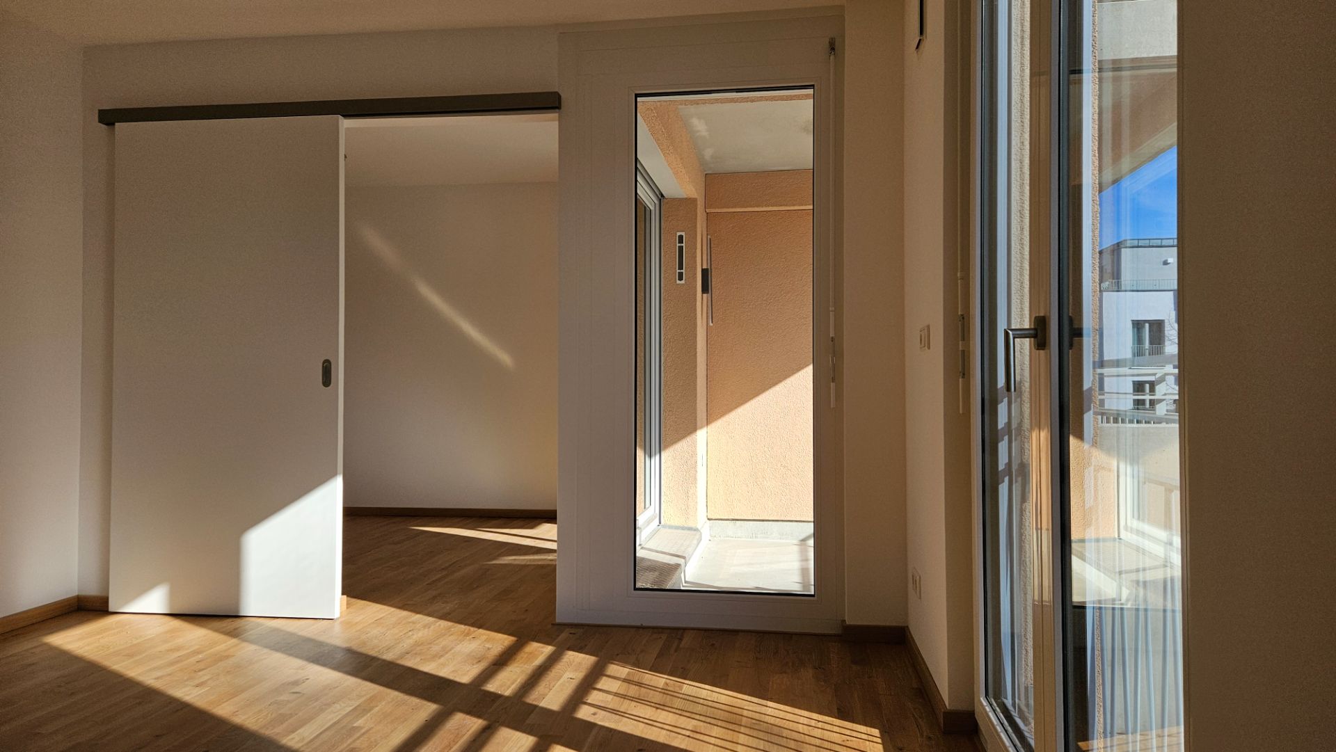 Heller Raum mit Holzboden, Schiebetür, Glasfront und Zugang zu einem Balkon.