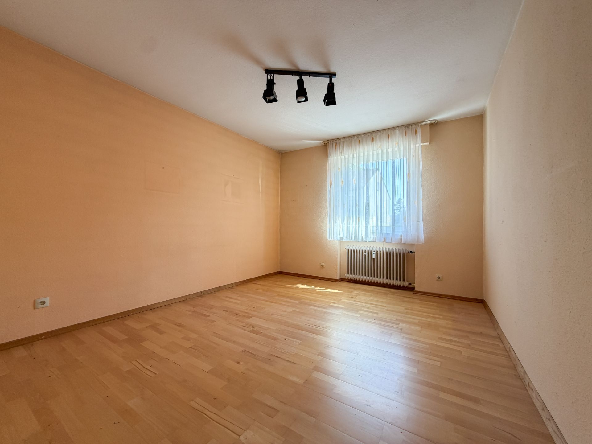 Leerer Raum mit hellem Holzfußboden, Fenster mit Vorhang und Deckenstrahler.
