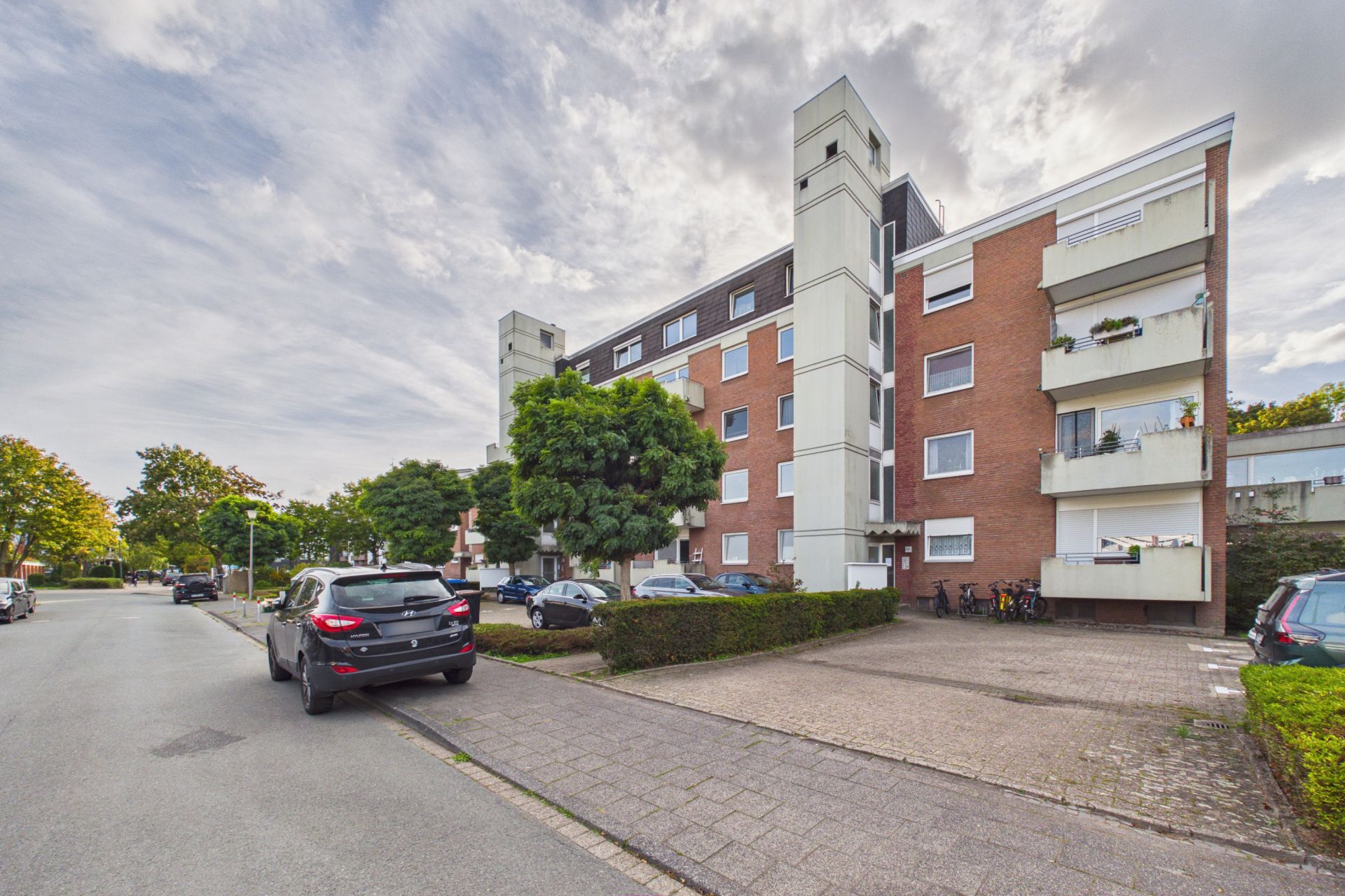 Mehrstöckiges Wohnhaus mit Balkonen, geparkten Autos und Bäumen an einer ruhigen Straße.