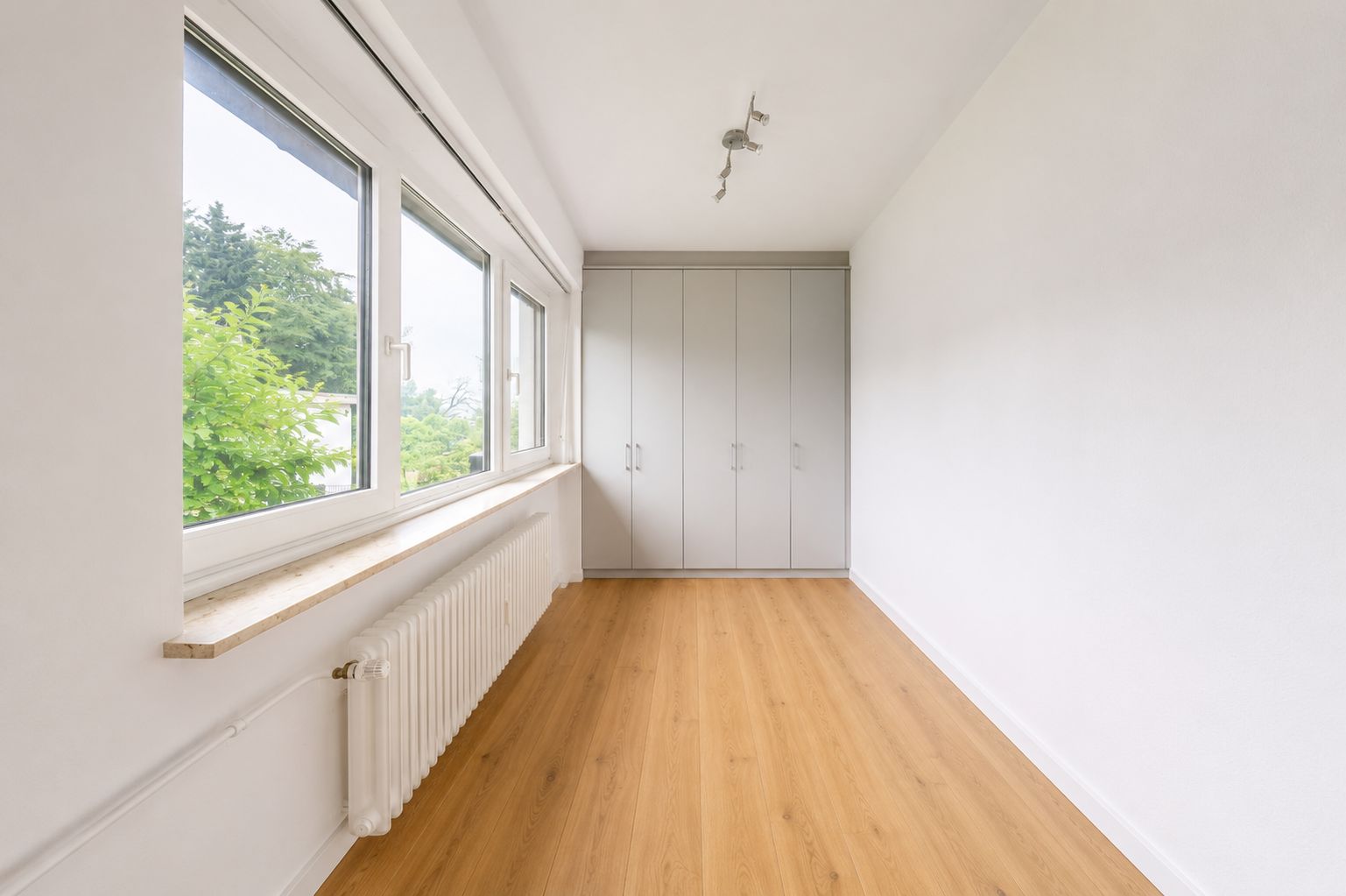 Heller Raum mit Holzfußboden, großen Fenstern, Heizkörper und Einbauschrank.