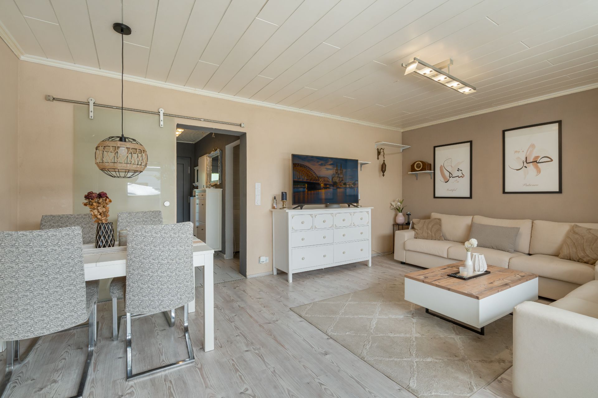 Helles Wohnzimmer mit Esstisch, Sofa, Kommode, Fernseher und Wandbildern.
