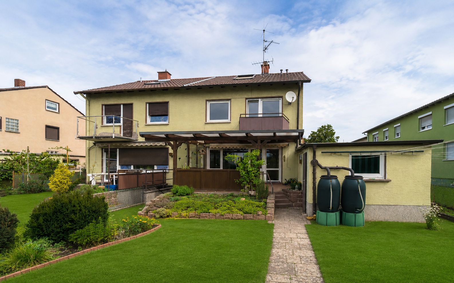 Zweistöckiges Wohnhaus mit Garten, überdachter Terrasse und Regentonnen im Hinterhof.