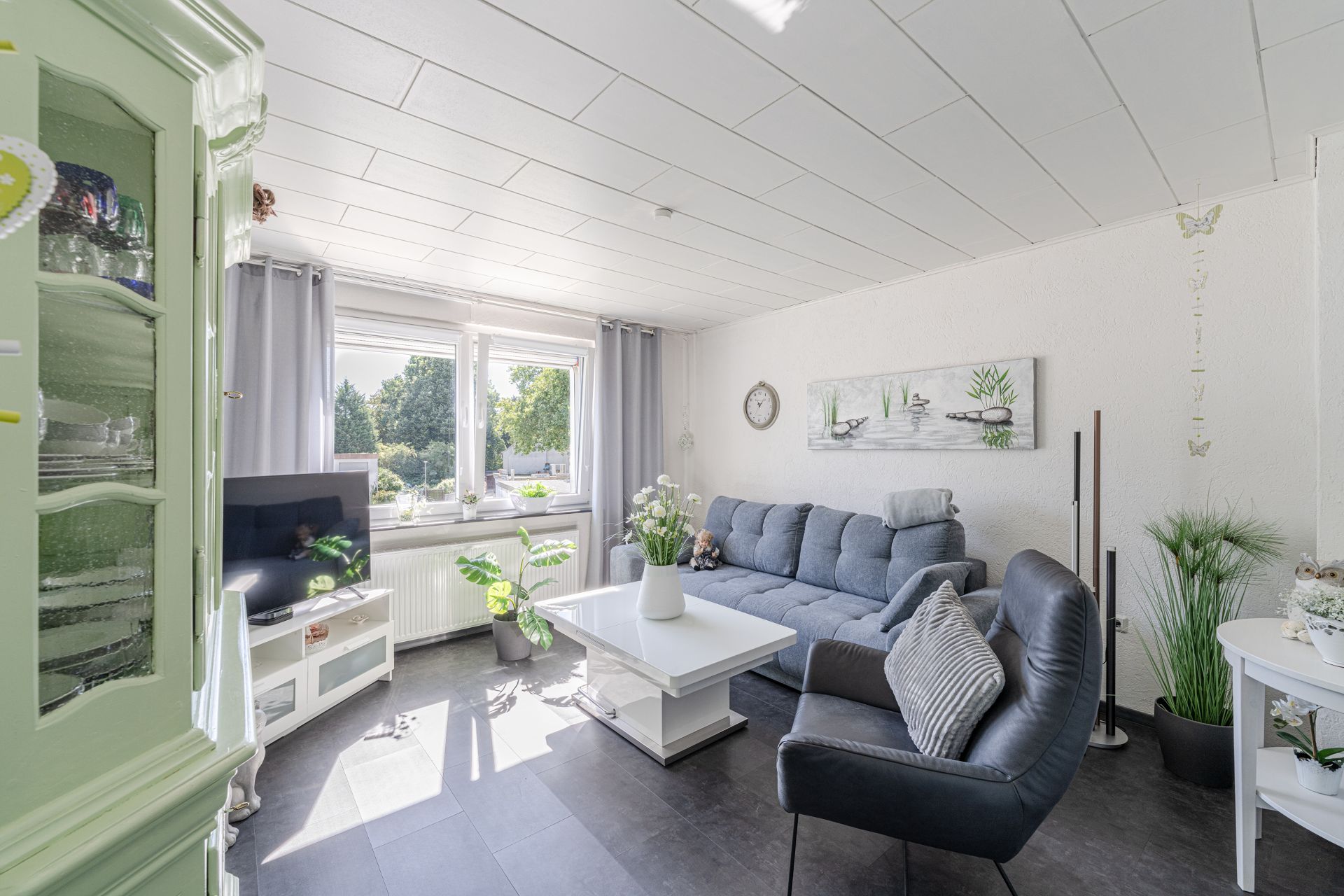 Helles Wohnzimmer mit grauem Sofa, Sessel, weißen Möbeln und großen Fenstern.